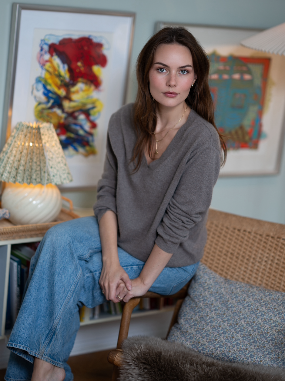 En ung kvinde med brunt hår i en grå sweater sidder på en stol i et hjem med kunstværker på væggen.