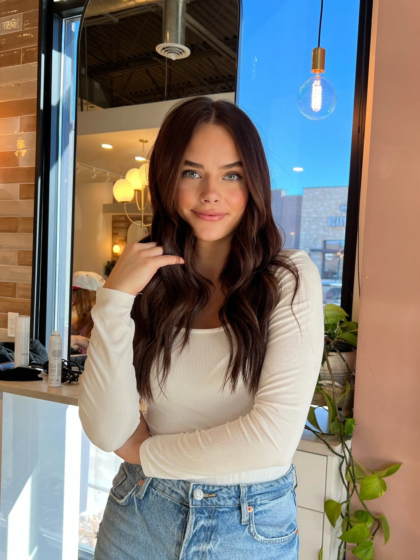 A young woman with long, wavy brown hair and blue eyes, smiling with a relaxed pose inside a modern cafe or restaurant.