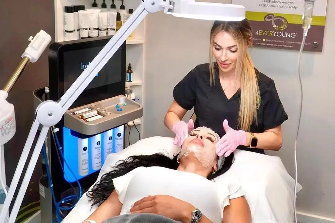 A woman lying on a treatment bed receiving a facial treatment at a spa or skincare clinic, with a skincare specialist applying a mask or product to her face.