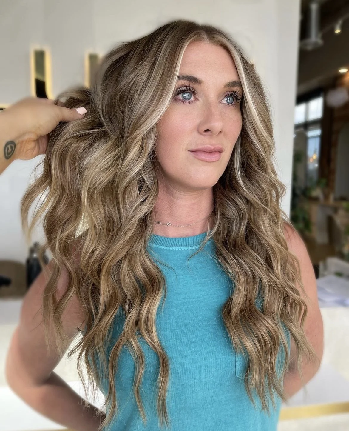 A woman with long, wavy blonde hair in a turquoise sleeveless top looks off to the side in a well-lit indoor space, while a hand adjusts her hair.