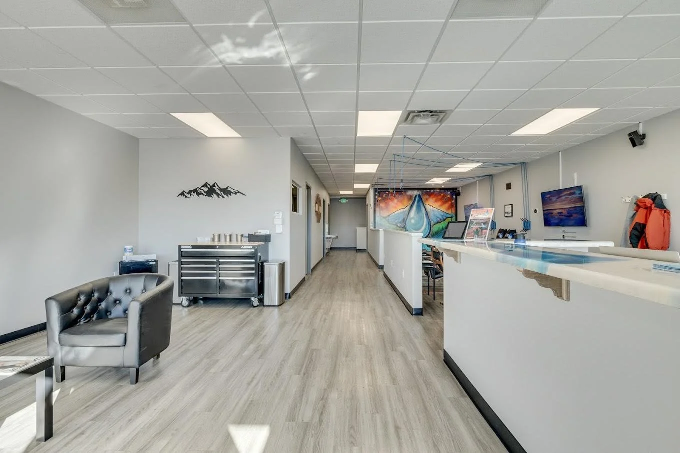 Empty office or reception area with a black leather armchair, a metal cart, and a long white counter with computers and a mounted television. Walls are light gray, with a mountain decal and a colorful mural of a mountain landscape.