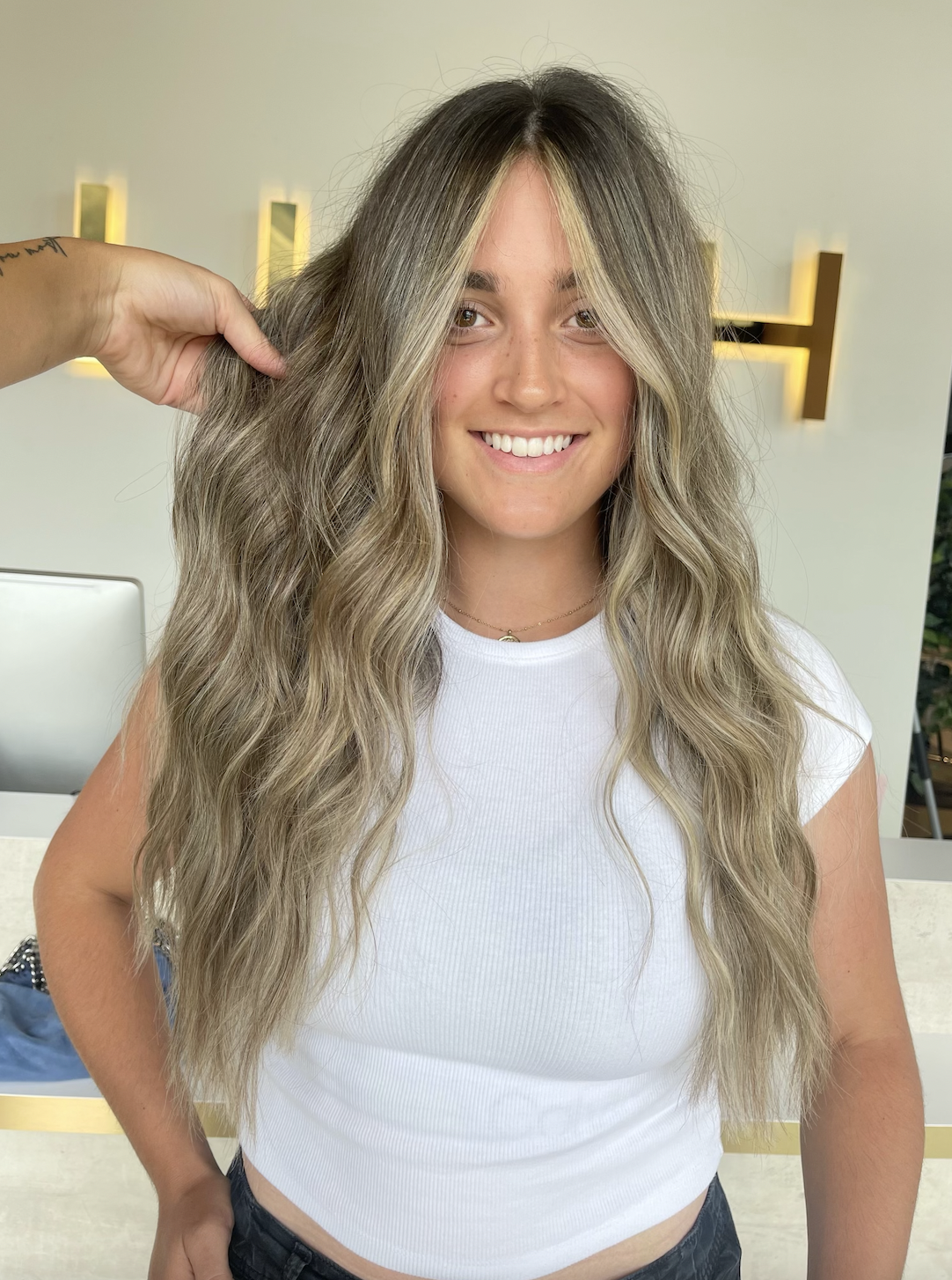 A woman with long, wavy blonde hair, wearing a white t-shirt, smiling at the camera, in a modern interior space.