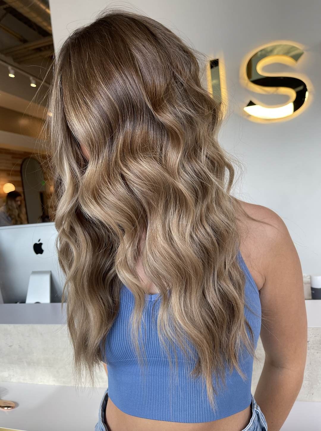 A woman with long, wavy blonde hair wearing a blue tank top standing in front of a modern interior with a large illuminated 'US' sign on the wall.