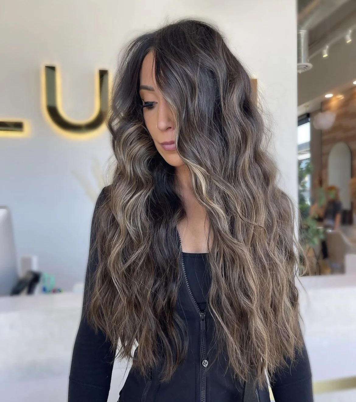 A woman with long, wavy hair that has dark roots and lighter ends, wearing a black zip-up top, standing inside a modern cafe.