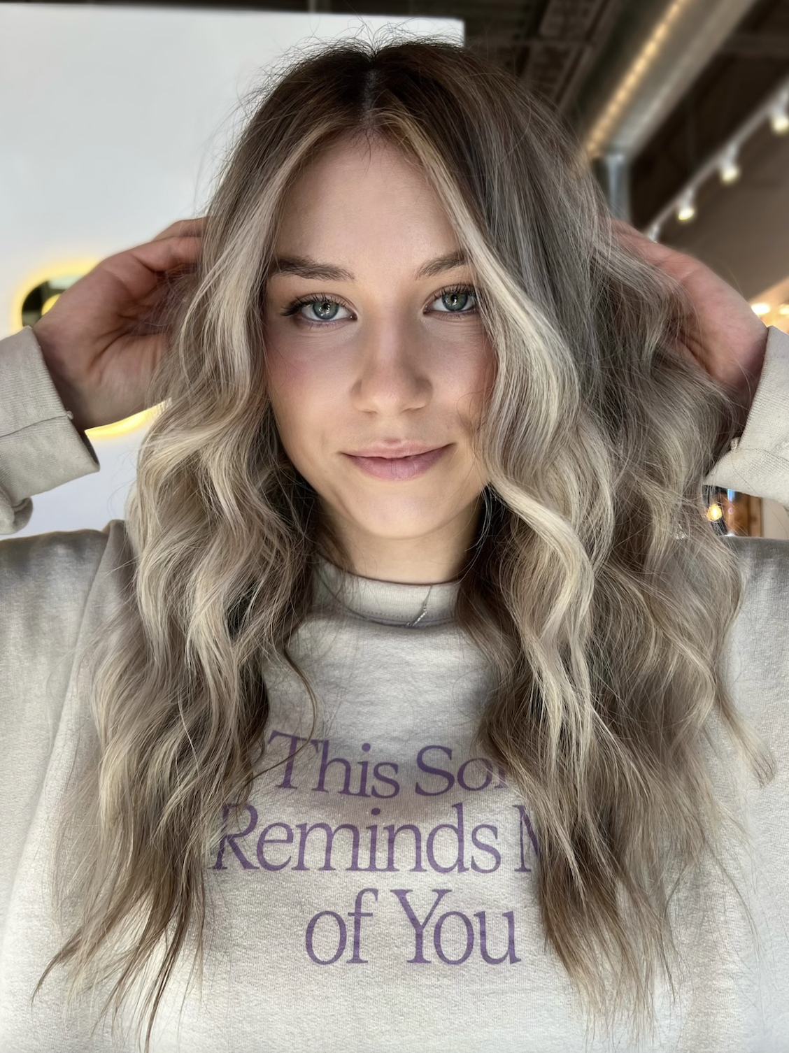 Young woman with wavy blonde hair and blue eyes, smiling, holding her hair back with both hands, wearing a light-colored shirt with purple text, in a well-lit, indoor setting.