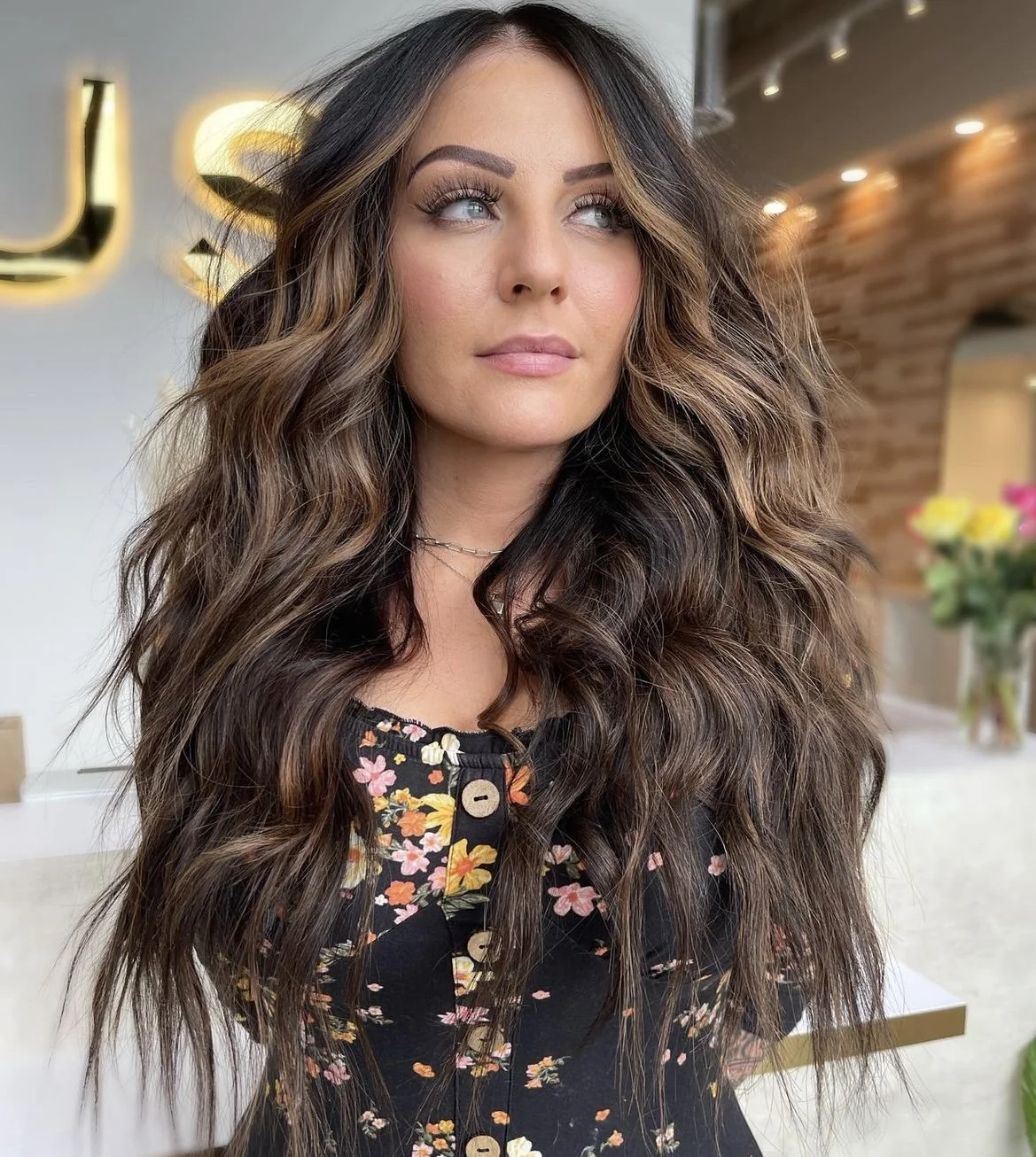 A woman with long, wavy brown hair and light makeup, wearing a black floral top, standing in a modern interior space with a blurred background.