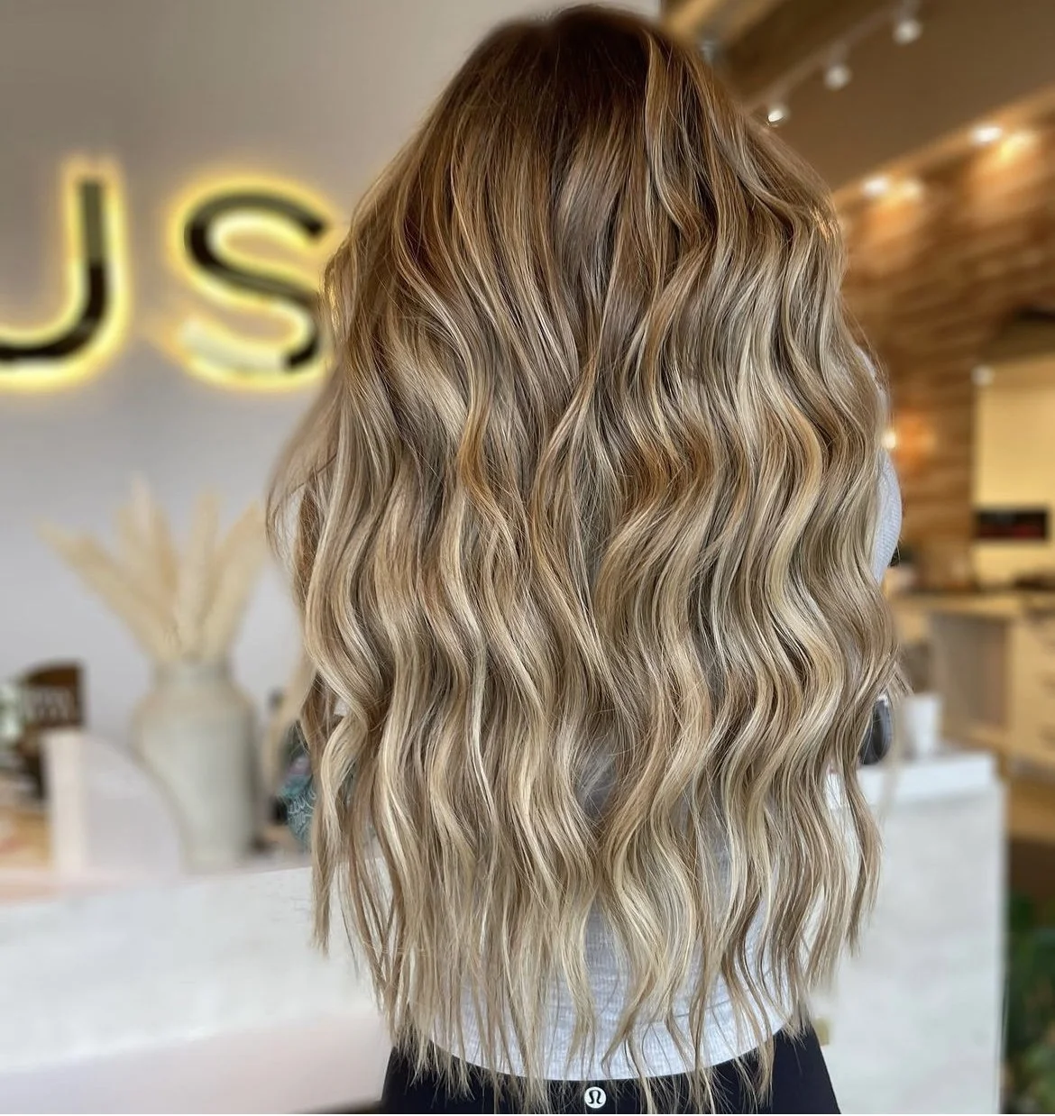 Back view of a woman with long, wavy blonde hair with darker roots, in a salon setting.