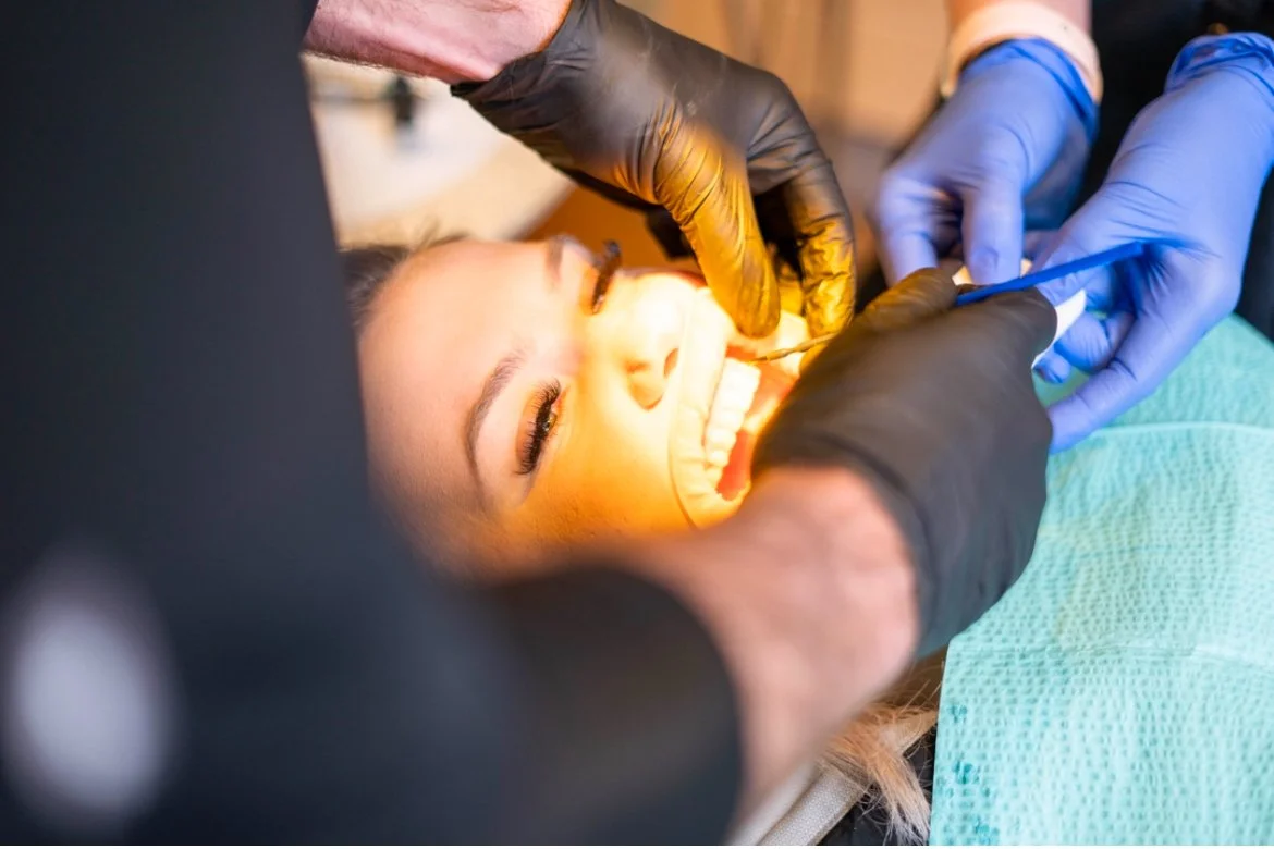 A woman receiving dental care, lying back in a dental chair with her mouth open, while dental professionals in gloves examine and work on her teeth.
