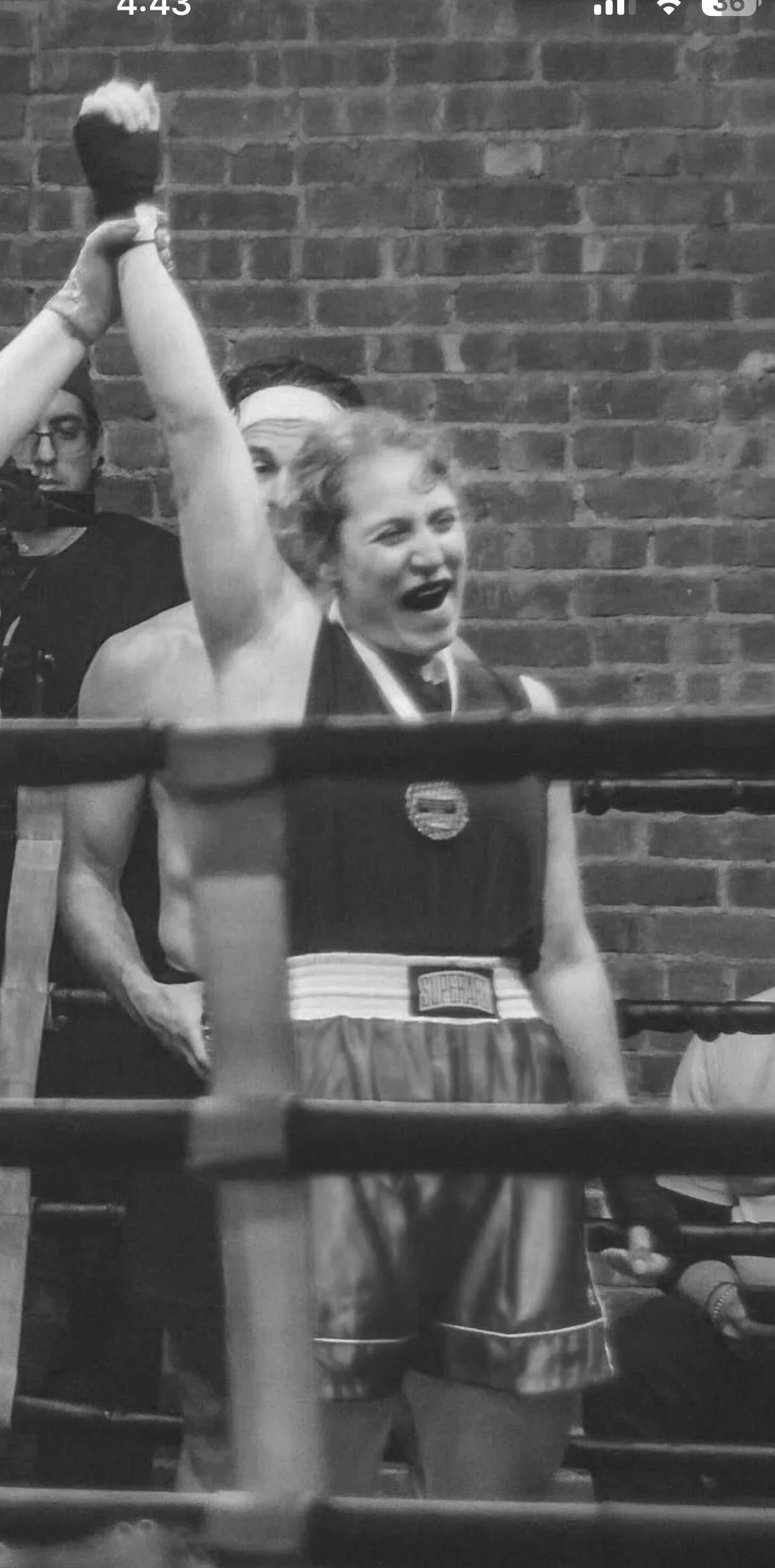 A young female boxer with short curly hair, wearing a black sports bra, blue boxing shorts, black mouthguard, and boxing gloves, raising her fist in victory or celebration inside a boxing ring with a brick wall background.