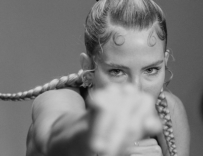 Close-up of Sydney with braided hair, looking intensely at the camera, with her hand raised in front of her face.