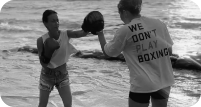 Sydney practicing boxing with another woman on a beach, wearing a shirt that says 'WE DON'T PLAY BOXING'.