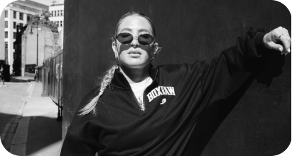 Sydney wearing sunglasses and a black jacket with a white logo, standing against a dark wall in an urban setting.