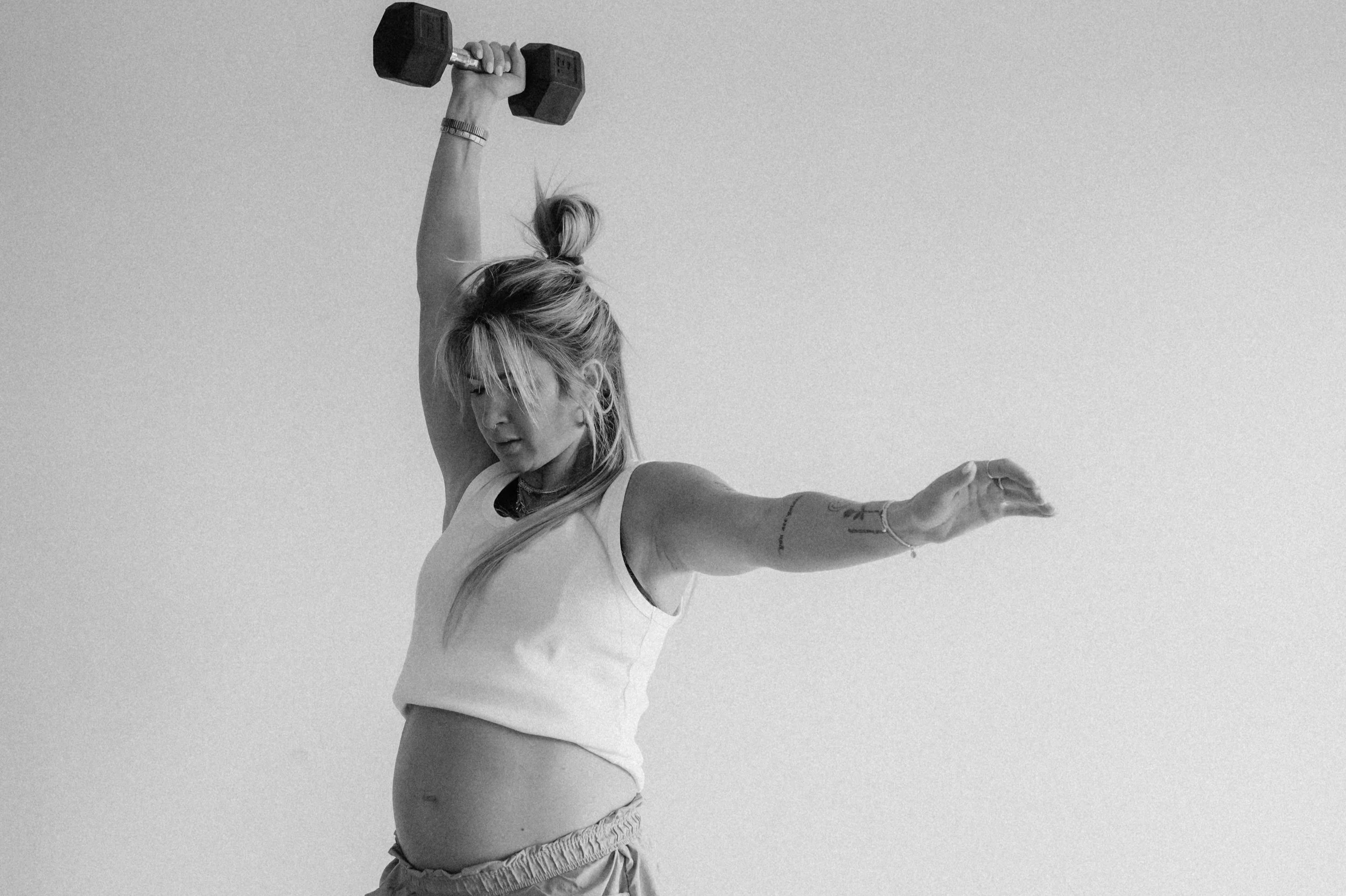 Sydney with long hair tied up, wearing a white tank top and loose shorts, holding a dumbbell in her right hand while balancing with her left arm extended outward, against a plain background.