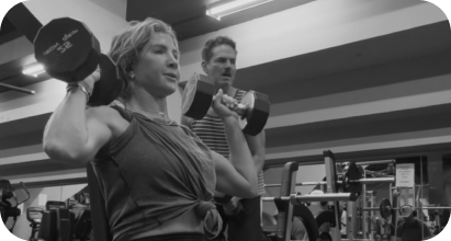 Sydney lifting a dumbbell with a man in the background at a gym.