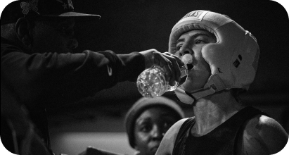 A person in a helmet drinks water from a water bottle held by another person. A woman in the background watches.