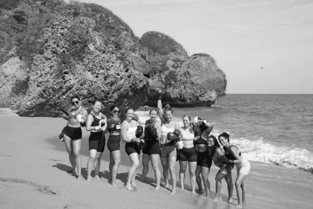 Group of nine women standing on a beach near large rocks, posing with boxing gloves and smiling.