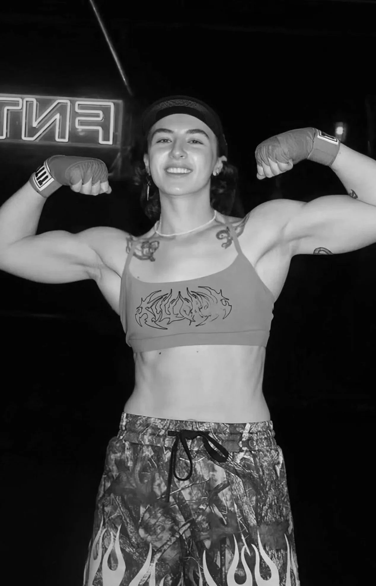 A young woman with tattoos, wearing a khaki sports bra and camouflage shorts, flexing her biceps with green boxing gloves in a dimly lit room with a neon red sign in the background.