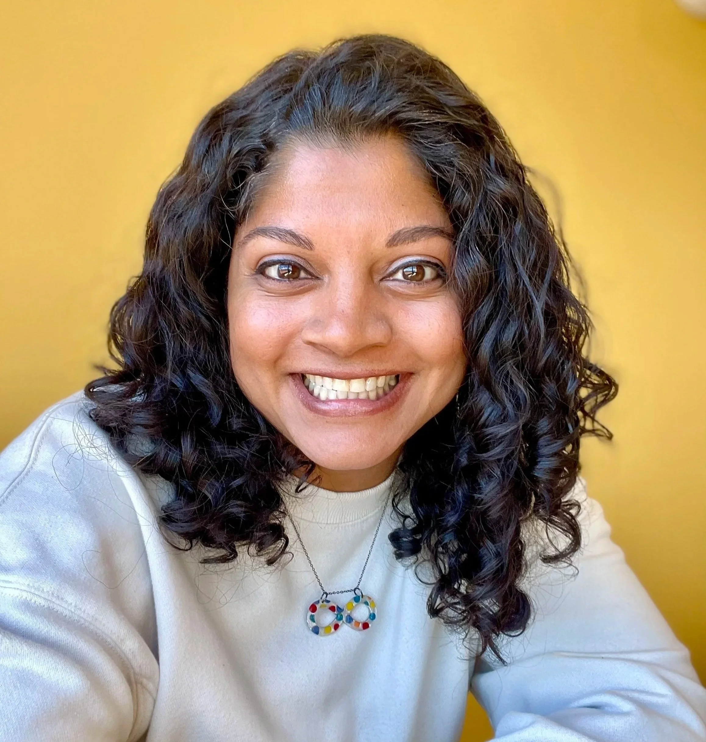 Close-up of a woman smiling, dark curly hair, wearing a white shirt and a colorful infinity necklace, against a yellow background.