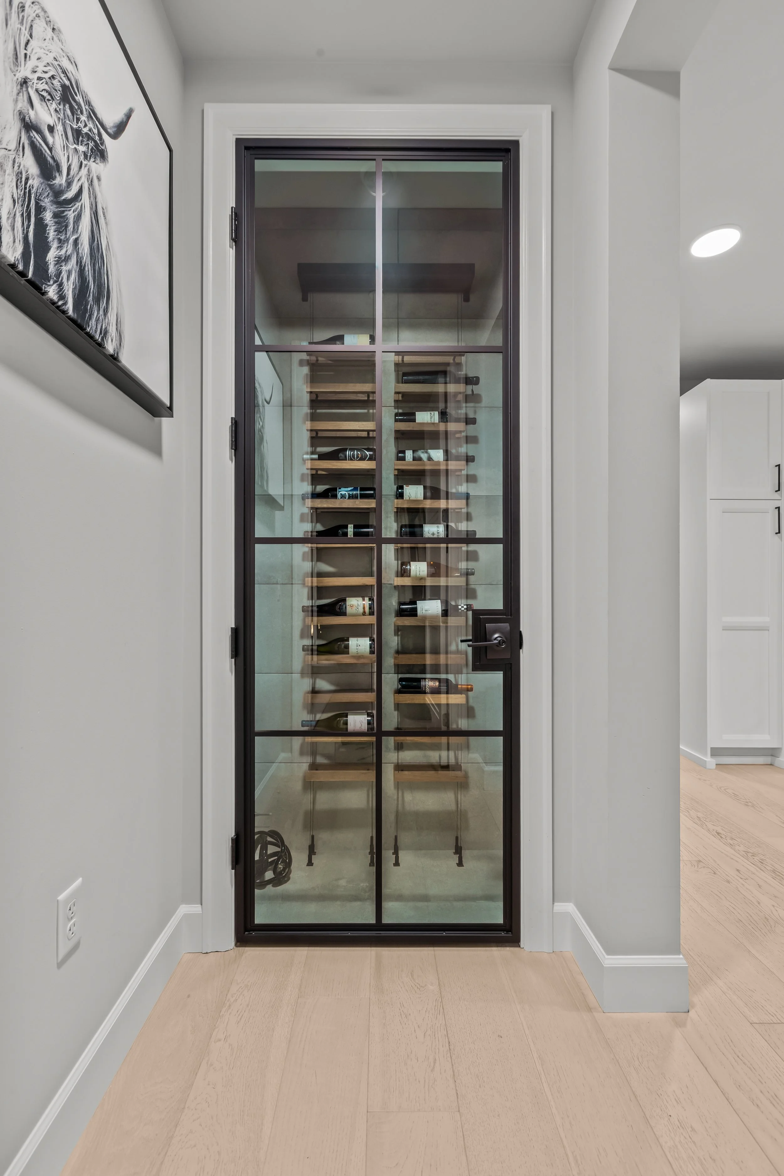 A glass door wine cellar closet with wooden shelves holding multiple bottles of wine, located in a modern home interior.