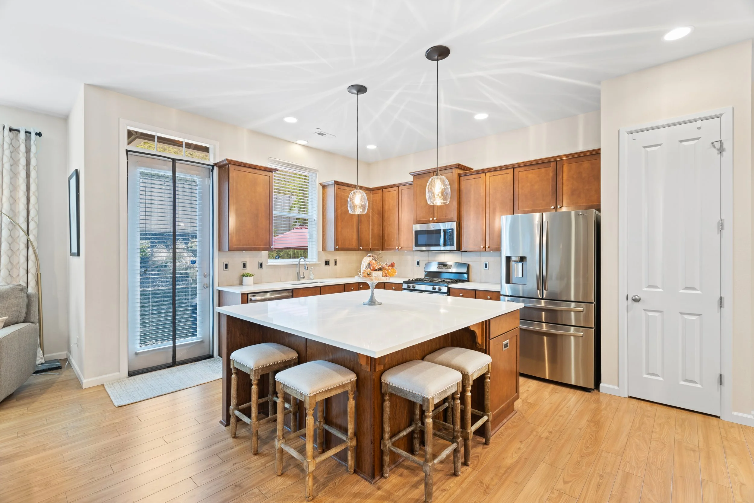 Modern kitchen with wooden cabinets, stainless steel refrigerator, island with white countertop, three barstools, and pendant lighting.