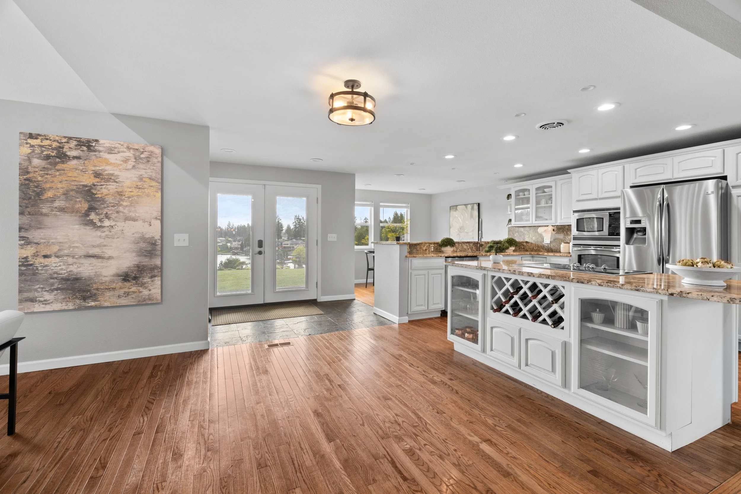 Bright kitchen with white cabinets, granite countertops, stainless steel appliances, and hardwood floors, leading to a deck with glass doors and a view of a neighborhood over a lawn.