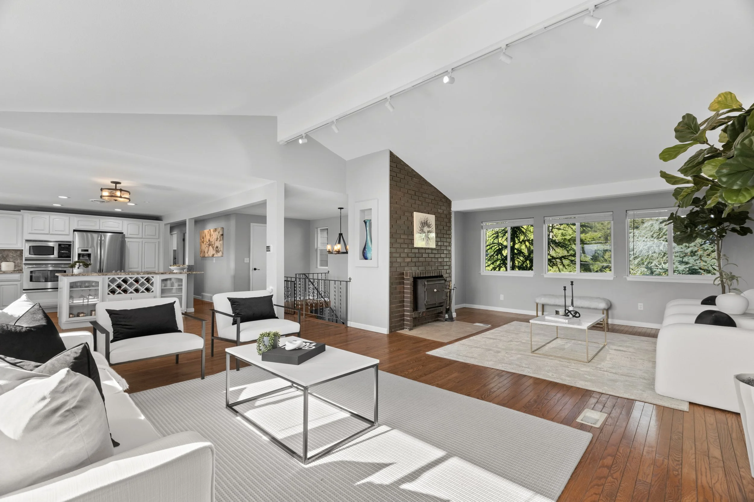 Spacious living room with white and black furniture, a brick fireplace, large windows, and hardwood floors.