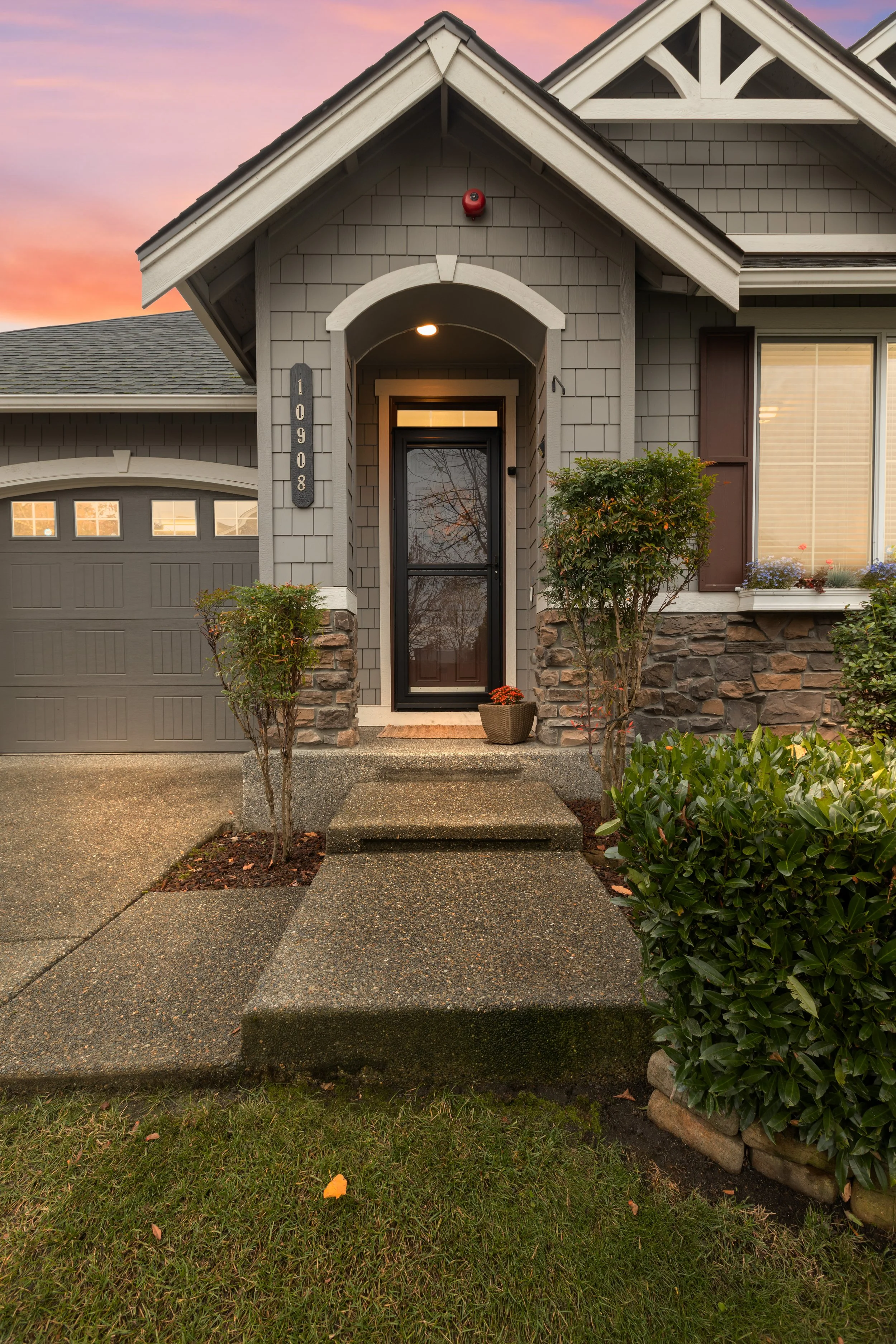 Front entrance of a house with a black door, concrete steps, shrubs, a potted plant, and a garage door on the side; during sunset.