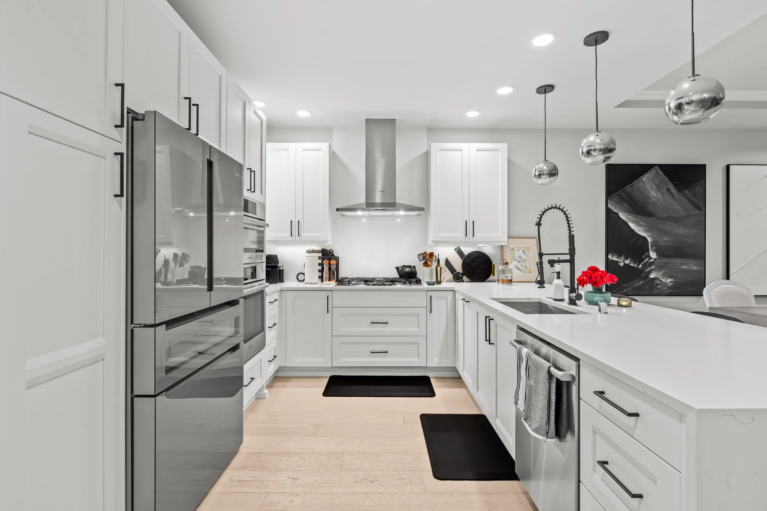 Modern kitchen with white cabinetry, stainless steel appliances, black backsplash, and pendant lighting, featuring a black faucet and a vase of red flowers.