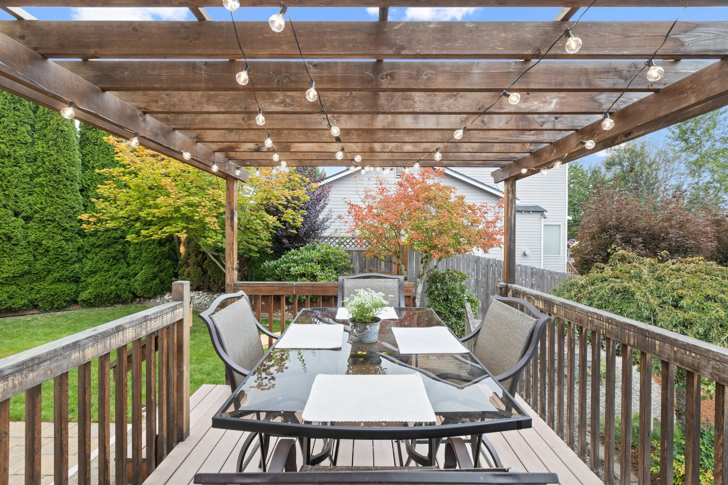 A backyard deck with a glass table and six chairs under a wooden pergola stringed with lights. The background features trees with green and red leaves and a white house.
