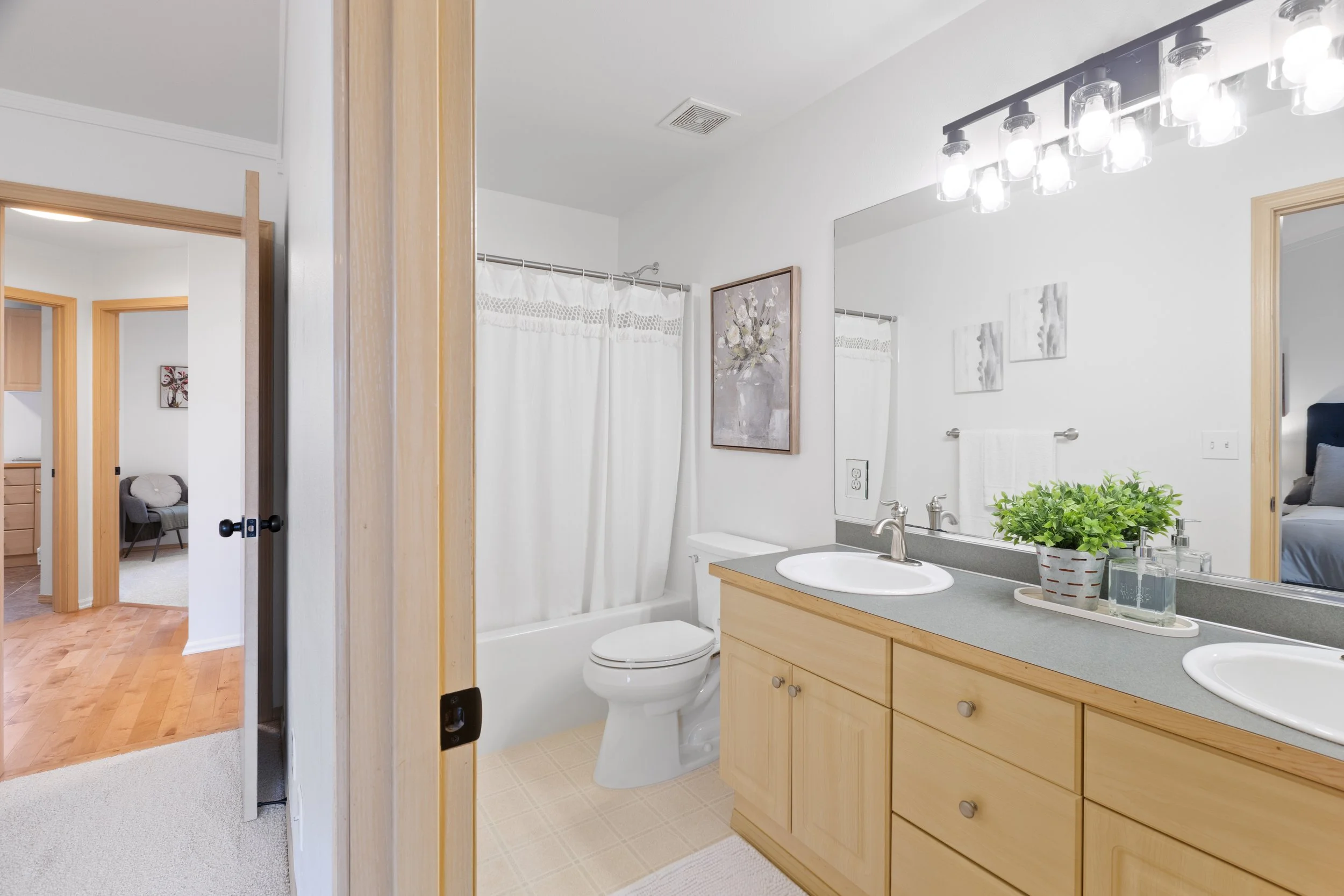 Bathroom with dual sinks, a large mirror, a plant on the counter, a shower with a white curtain, and artwork on the wall. Adjacent to a bedroom with a bed visible.