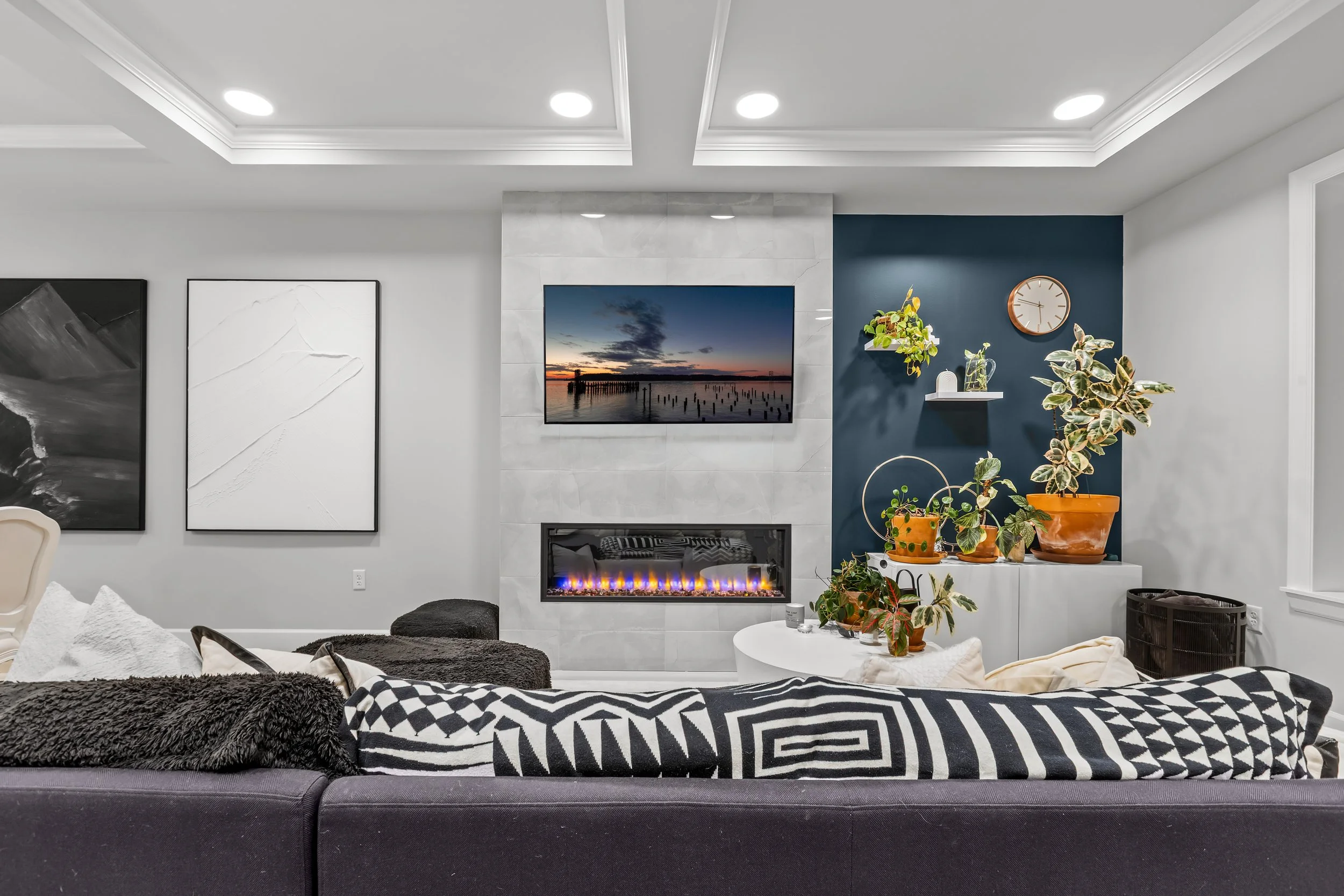 Modern living room with a black sofa, patterned pillows, a wall-mounted flat-screen TV showing a sunset over a pier, a white fireplace, and a blue accent wall with plants on white shelves.
