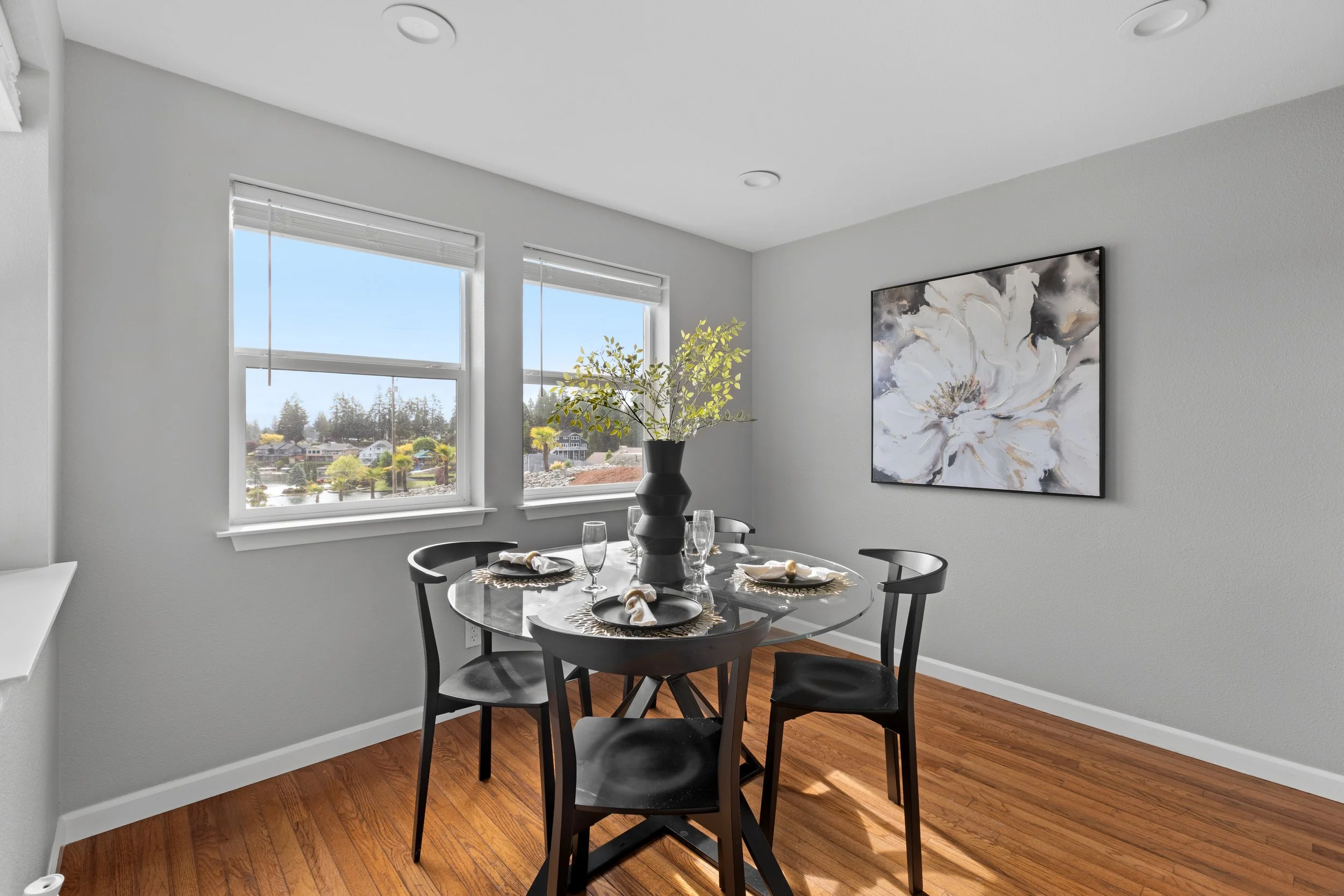 Dining area with a round glass table set for four, a tall black vase with green leafy branches, and a large floral painting on the light gray wall, with two windows showing a view of houses and trees outside.