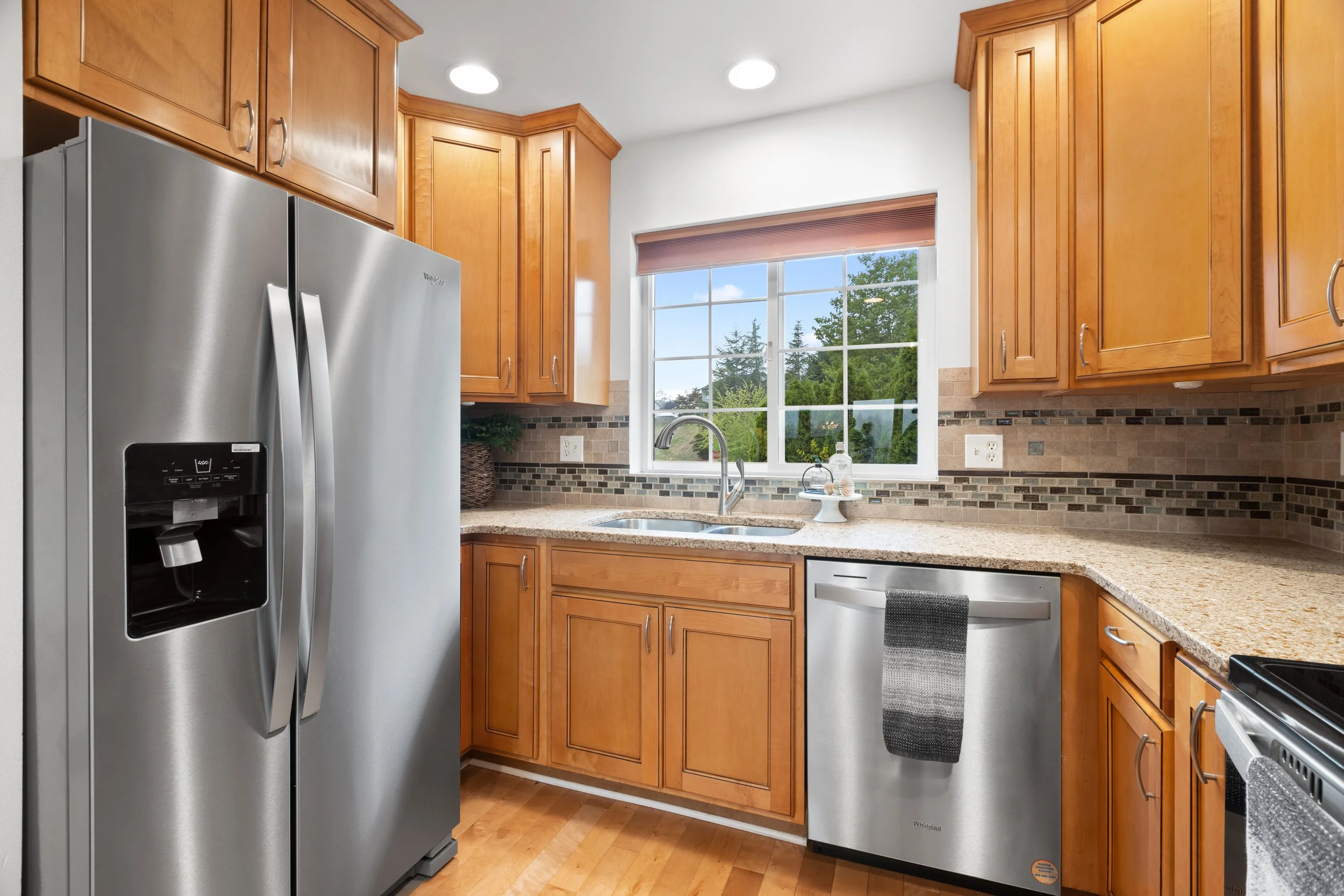 Kitchen with wooden cabinets, stainless steel refrigerator, dishwasher, and stove. A window above the sink showing trees and blue sky.