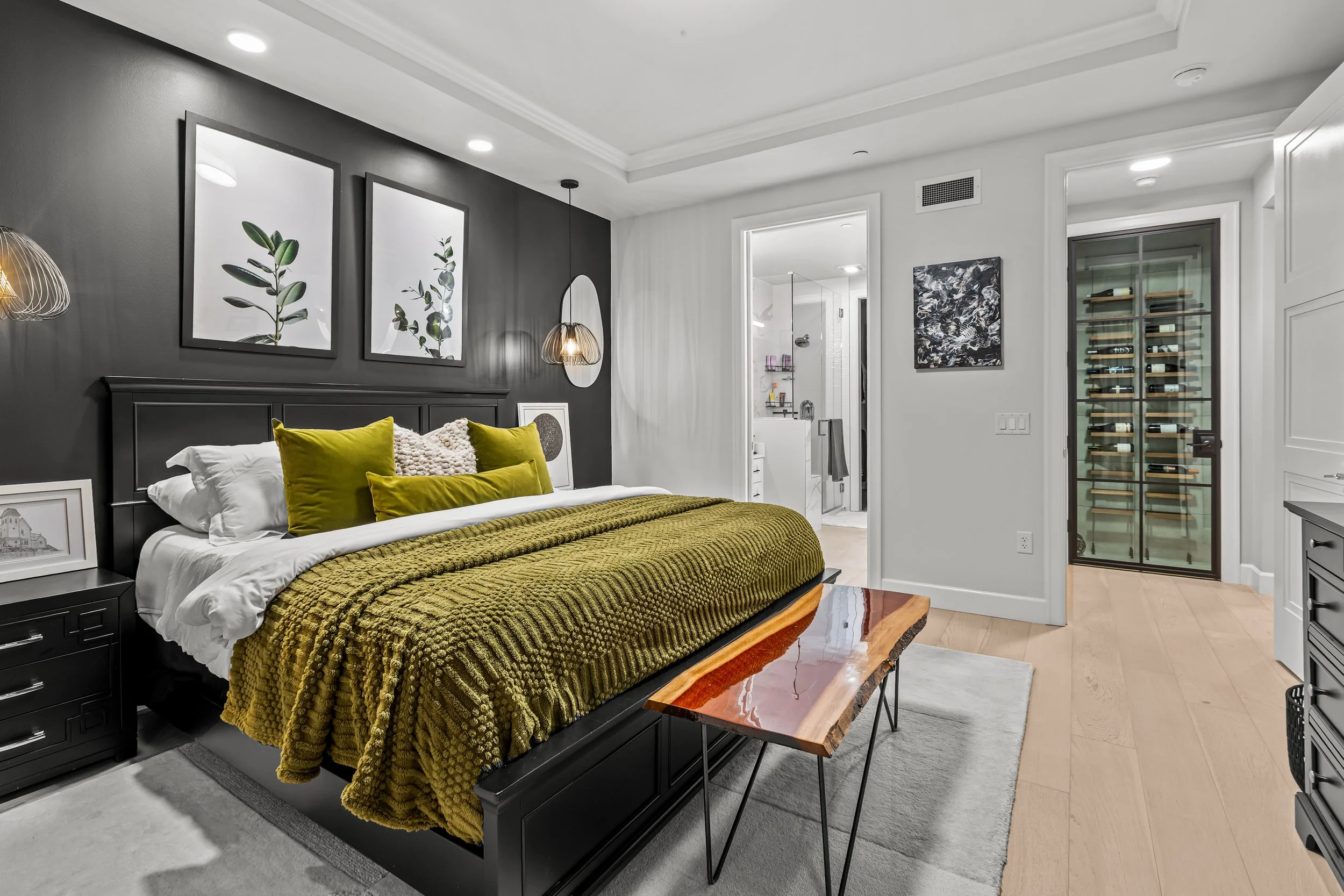 Modern bedroom with black accent wall, green bedding, and minimalist decor, including framed botanical art, pendant lighting, and a wood bench, with an en suite bathroom and a glass wine storage cabinet.