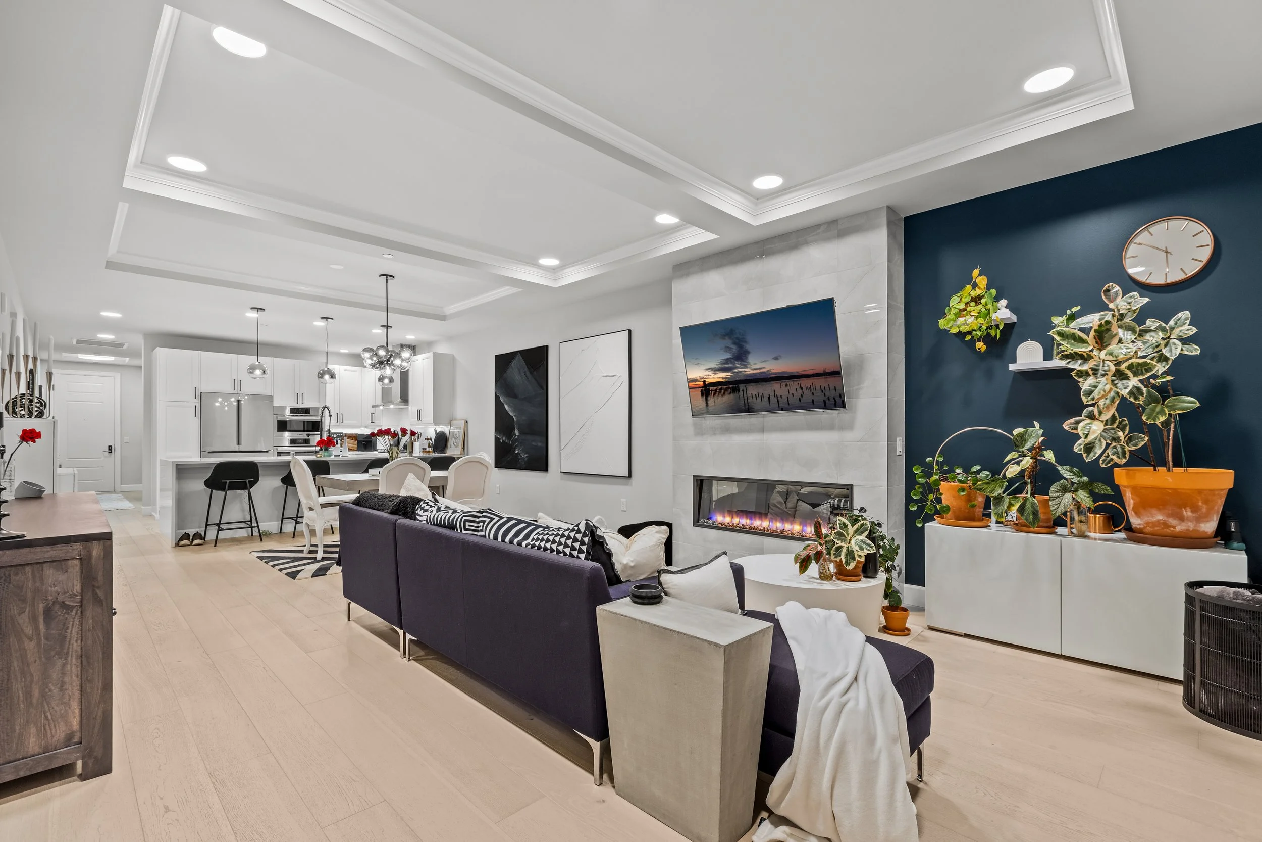 Living room with fireplace, wall-mounted TV, and large potted plants, opening to a kitchen and dining area with pendant lighting and artwork.