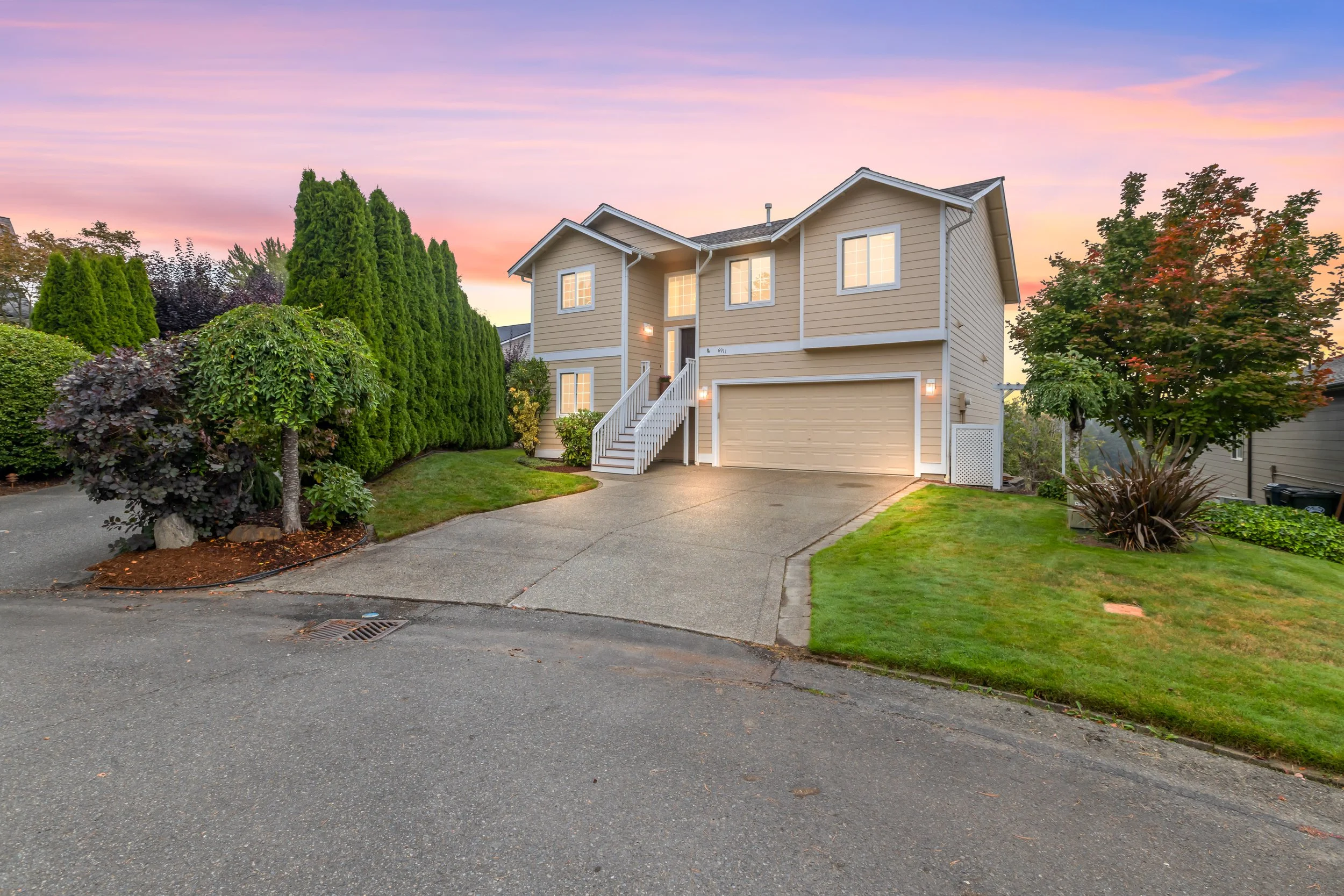 A two-story beige house with white trim, a two-car garage, and steps leading to the front door, with landscaped green lawns, trees, shrubs, and a pink sky at sunset.