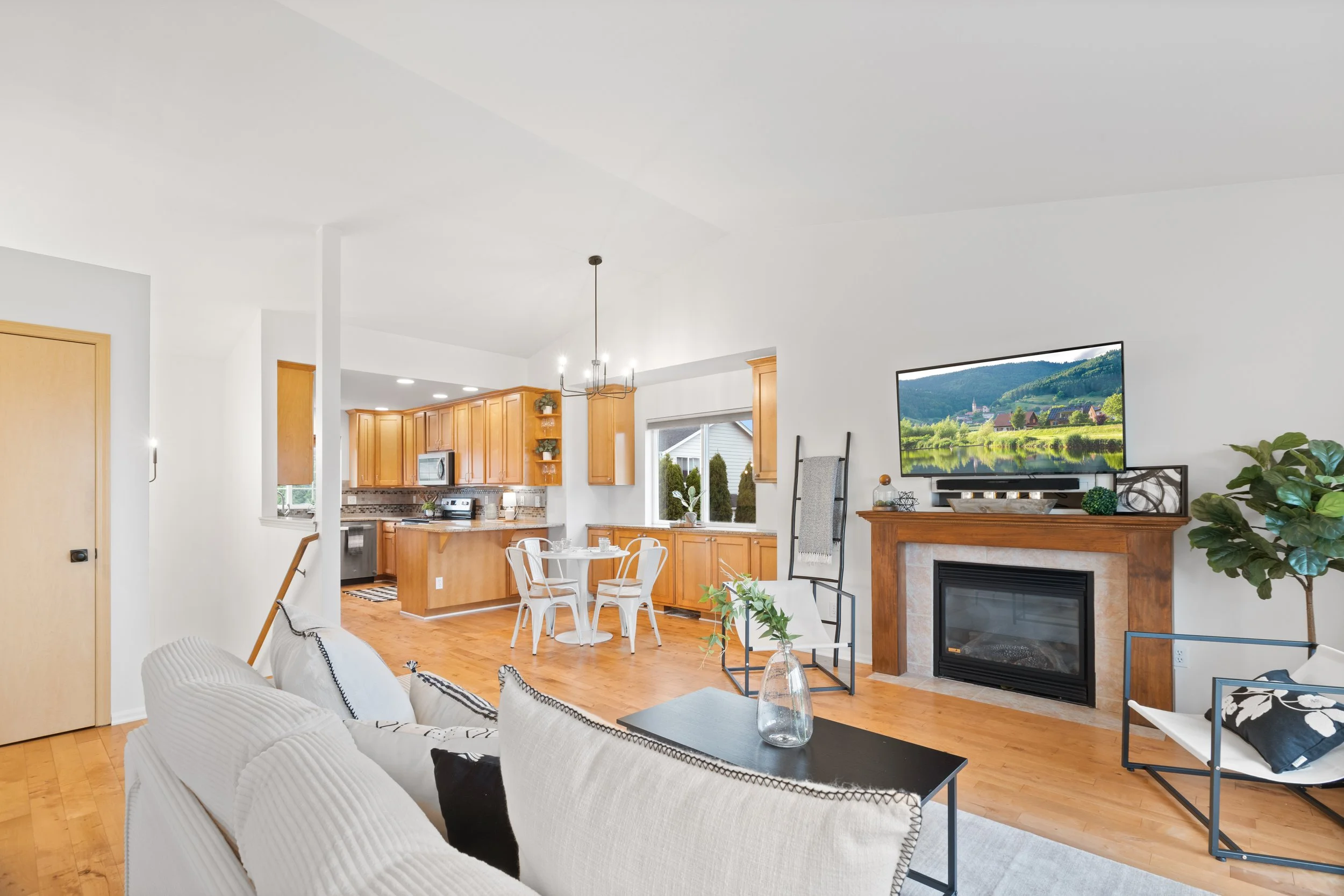 Living room with a fireplace, flat-screen TV, white couch, armchair, glass coffee table with a vase, and a large window. Adjacent dining area and kitchen with wooden cabinets, microwave, and stainless steel appliances.