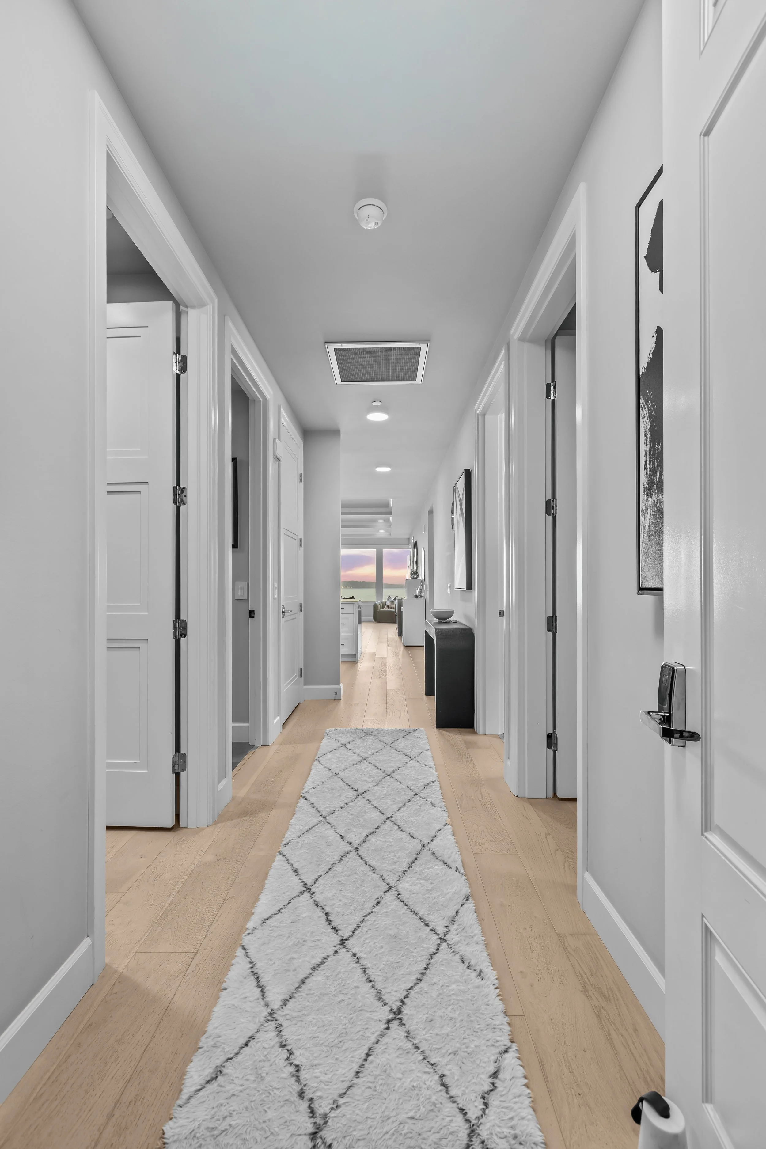 A hallway in a modern home with white walls, light wood flooring, a white fluffy rug, and multiple closed white doors, leading to a living room with large windows showing a sunset sky.