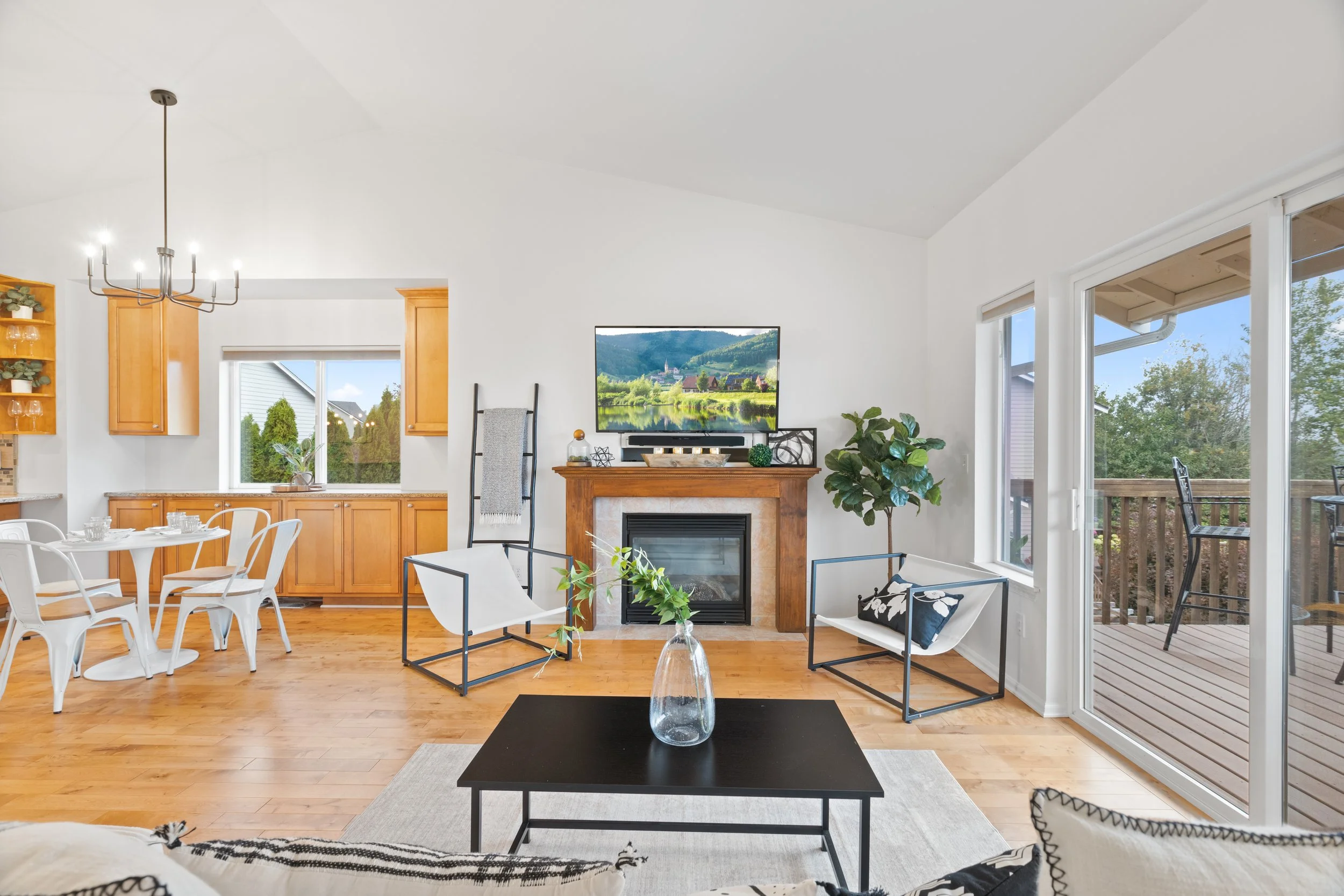 Bright living room with a fireplace, flat-screen TV, black coffee table with a glass vase and green plant, white and black chairs, a large sliding glass door leading to a wooden deck, and a view of trees outside.