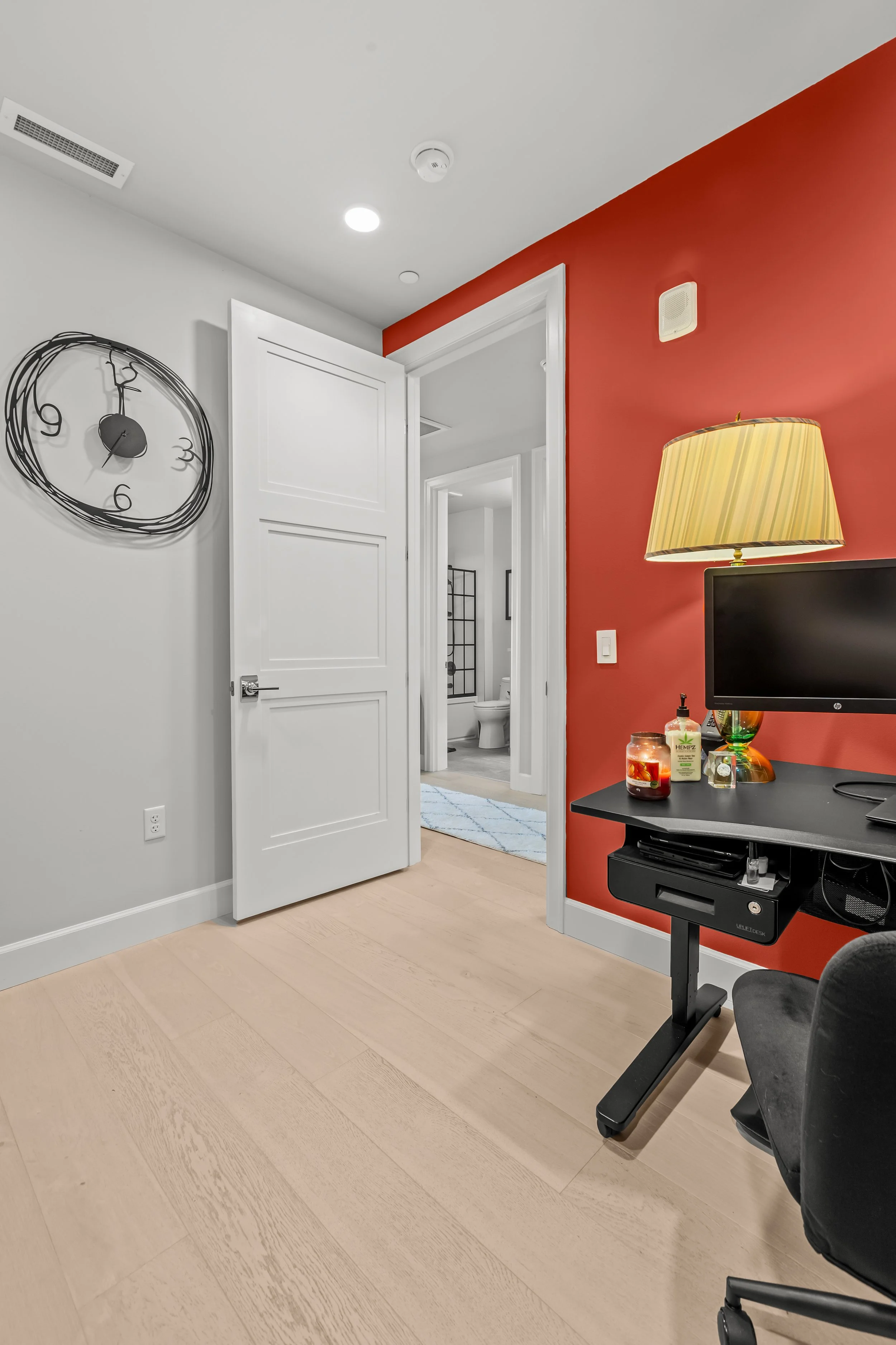 Interior of a modern living space showing a room with a white door open to a bathroom with a toilet and shower. The room has a red accent wall, a black desk with a computer monitor, a table lamp with a yellow shade, and various small items on the des