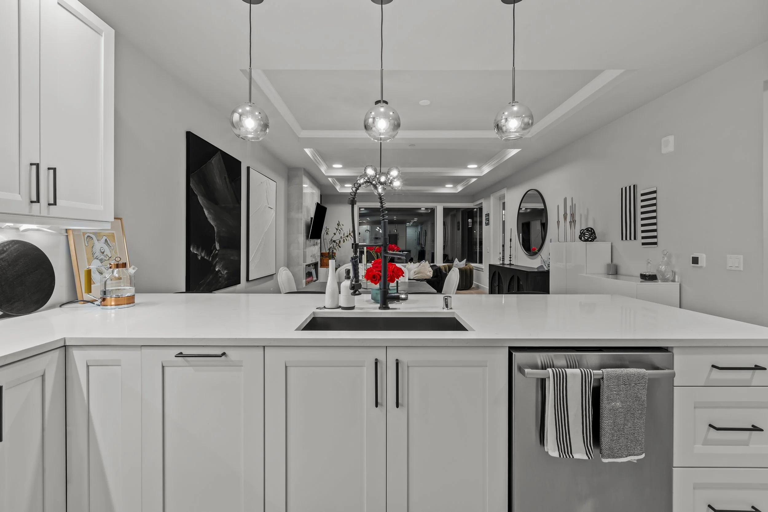 Modern kitchen with white cabinets, a white countertop, a black sink, and a chrome faucet. Decorative items include vases, art pieces, and a striped towel hanging on the dishwasher. Above, there are three spherical pendant lights. The kitchen opens i