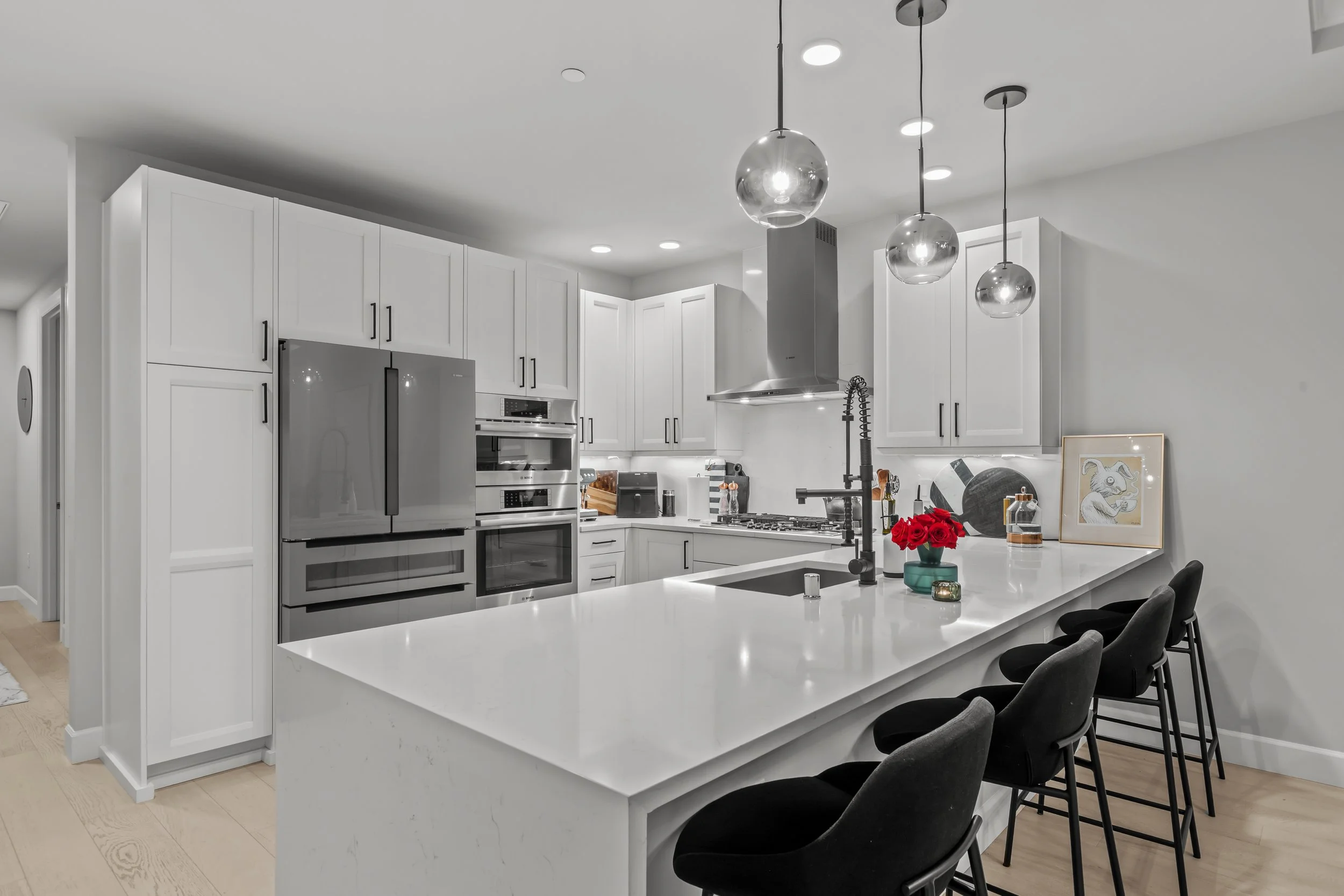 Modern kitchen with white cabinets, stainless steel appliances, a central island with a white countertop, black bar stools, and pendant lights.