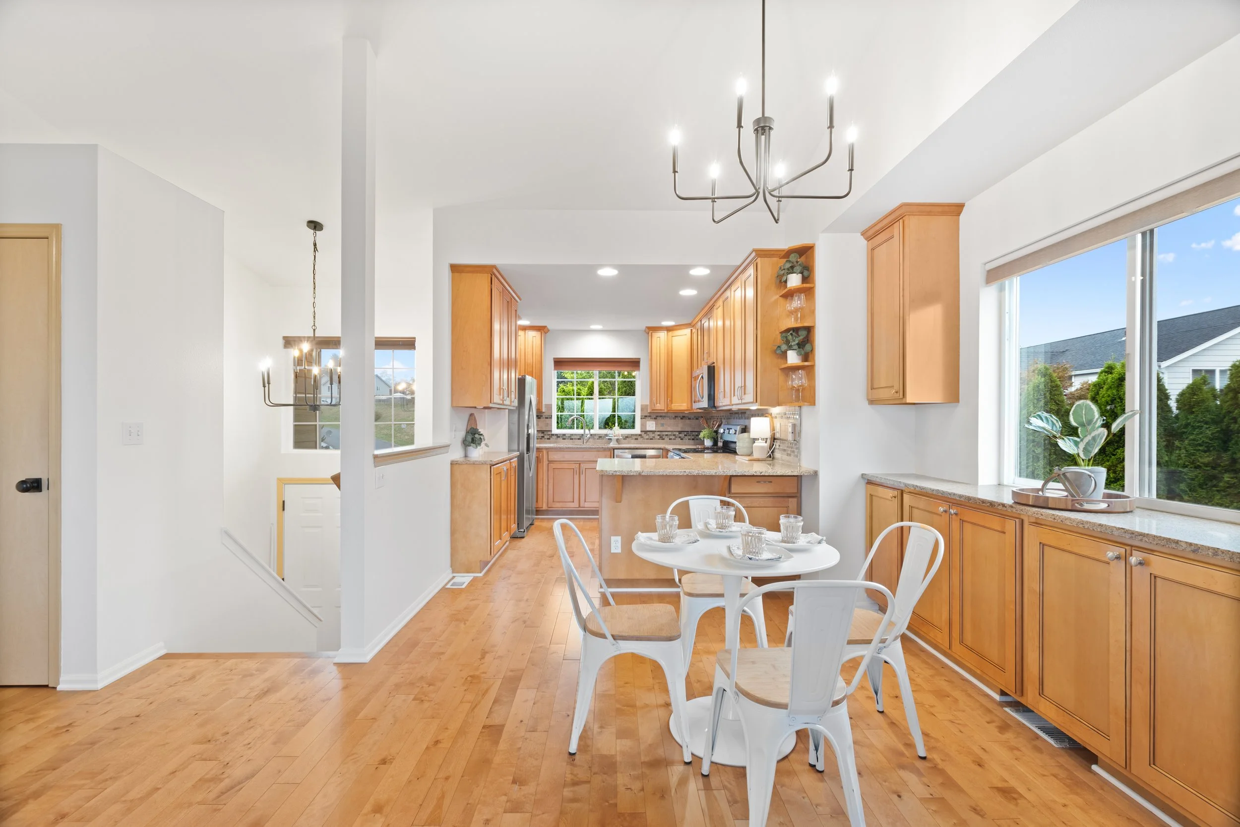 View of a bright kitchen with wooden cabinets, large windows, a small white dining table with four chairs, and a modern chandelier.