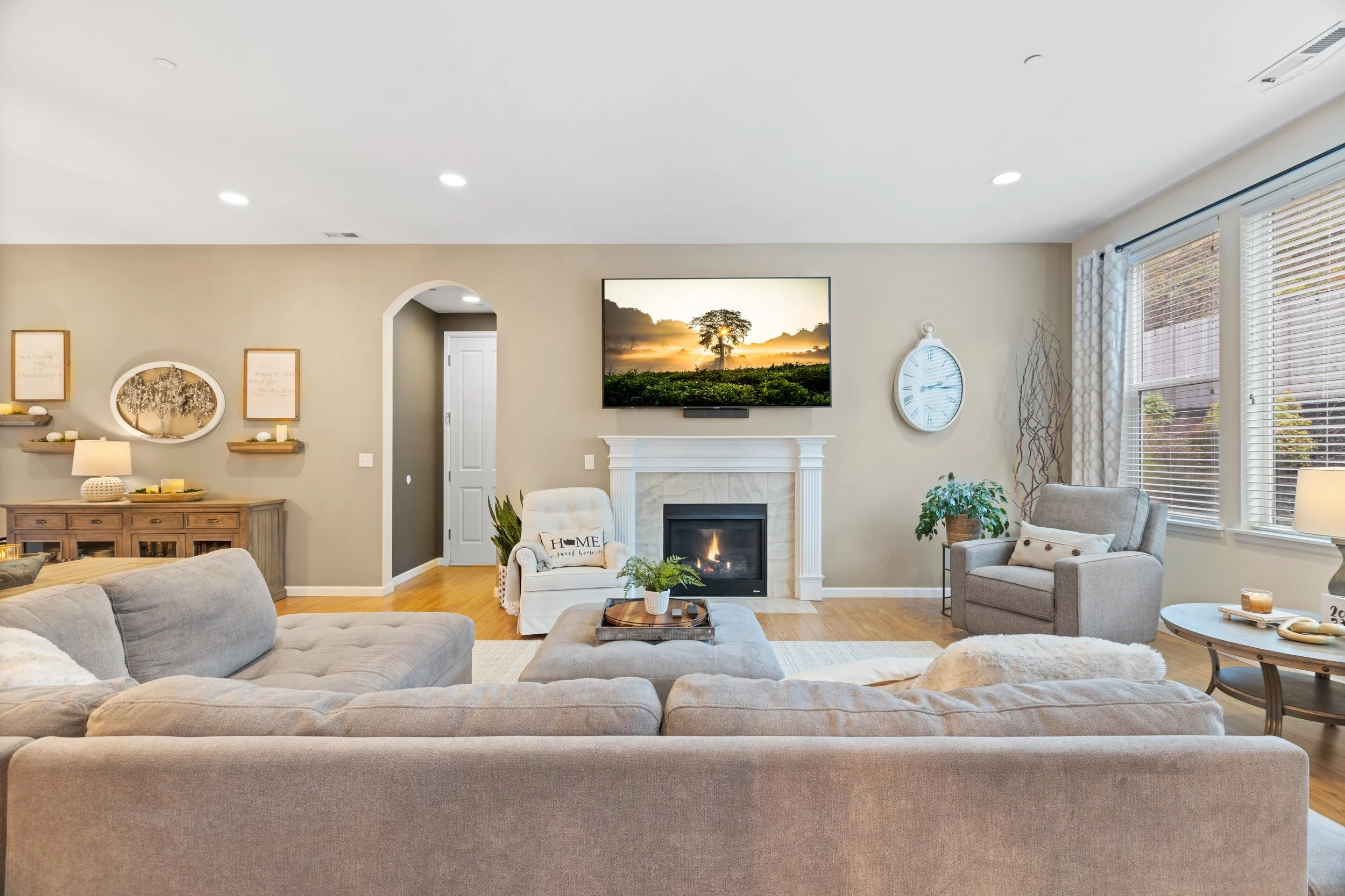 Living room with beige walls, a white fireplace, a flat-screen TV above it showing a sunset scene, a beige sectional sofa, two armchairs, a coffee table, and large windows with blinds allowing natural light.