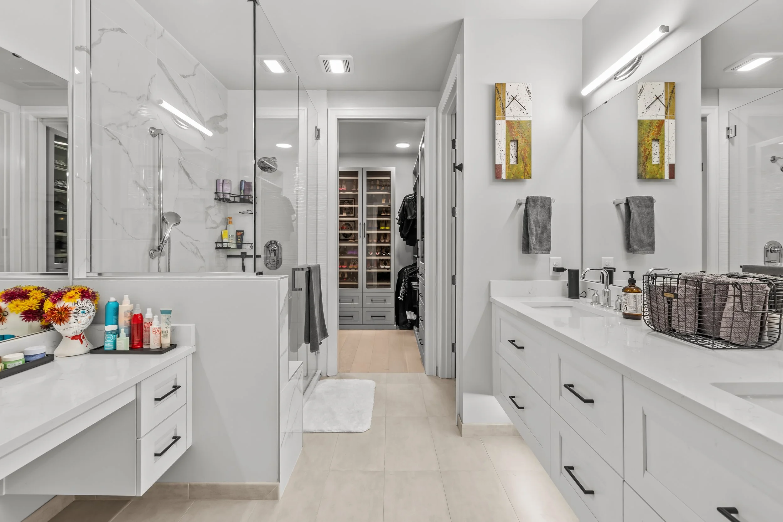 Modern white bathroom with a double vanity, large mirror, and a glass-enclosed shower. Visible shelves with toiletries and a walk-in closet with framed shoes in the background. Decorative artwork and gray towels on the wall.
