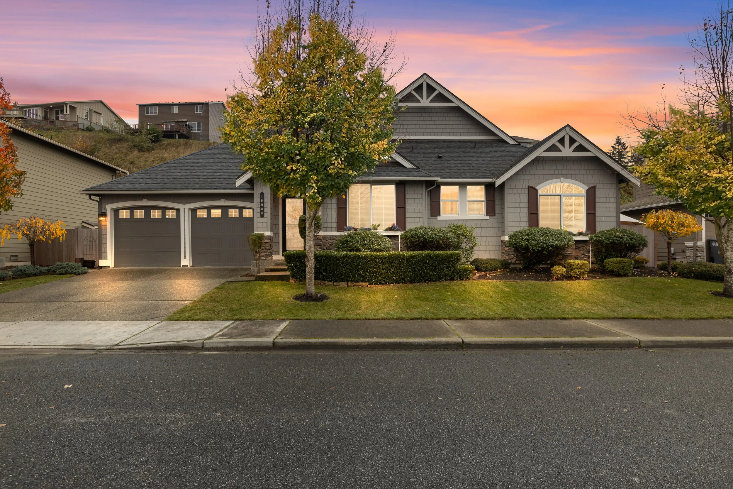 A modern suburban house during sunset with a manicured lawn, trees, and shrubs in front. The house has a double garage with a concrete driveway, and the sky shows shades of pink, purple, and orange.