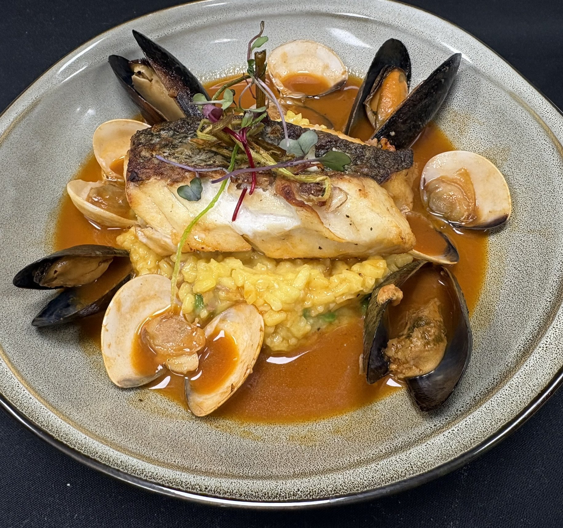 A plated seafood dish with grilled fish, mussels, and clams served over rice and surrounded by tomato-based sauce, garnished with microgreens.