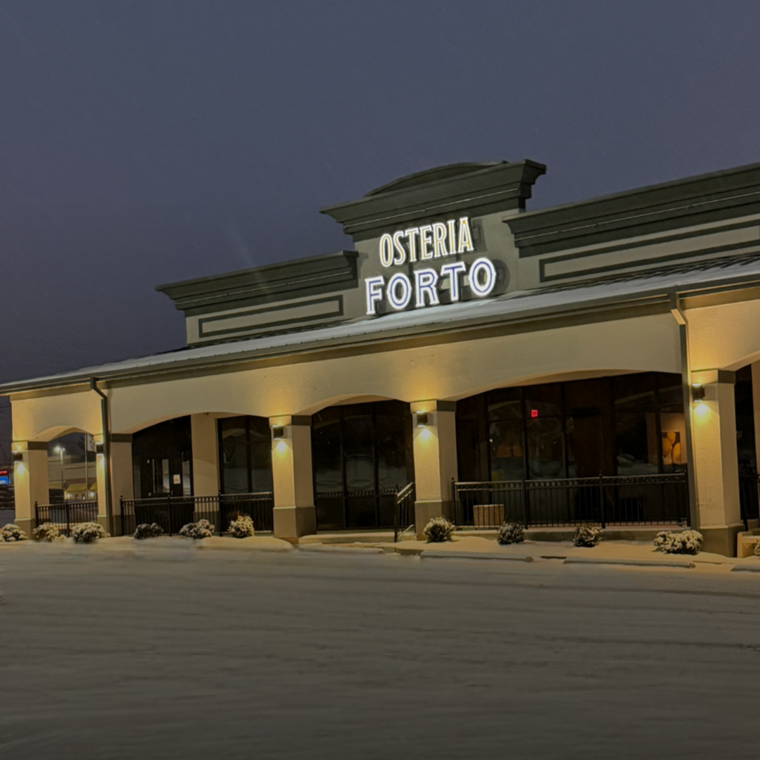 A restaurant named Osteria Forto illuminated at night, with a sign on the roof, snow on the ground, and some small bushes covered with snow outside.