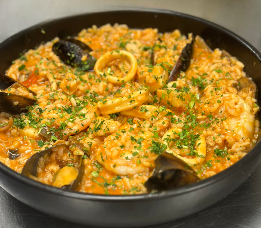 A black bowl filled with seafood paella containing mussels, calamari, and rice garnished with chopped parsley.
