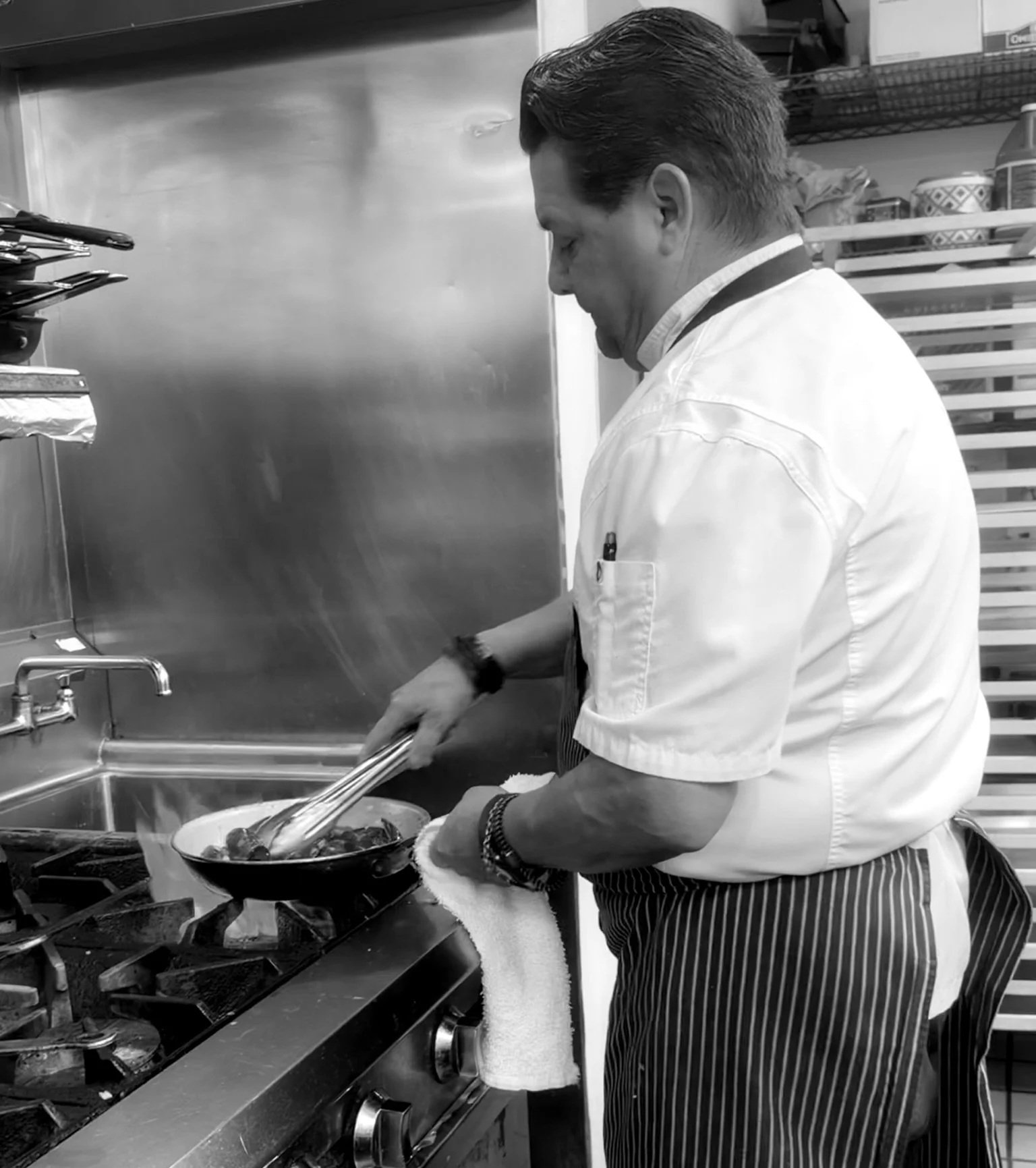 A male chef cooking on a stovetop in a professional kitchen, using tongs to handle food in a frying pan.